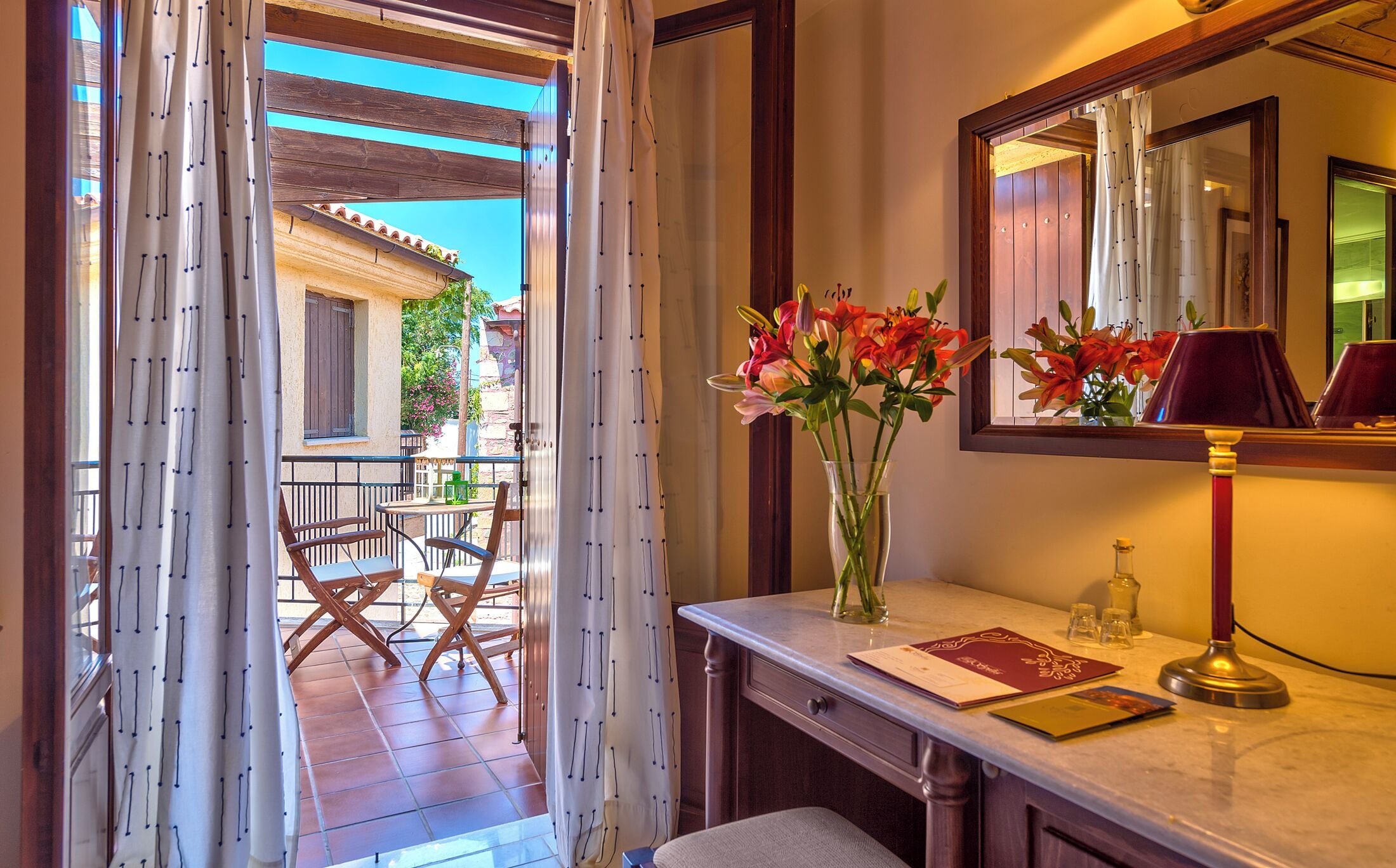 A hotel room with a balcony, a dining area with chairs, a vase with flowers, a mirror, and a desk with a lamp and a book.