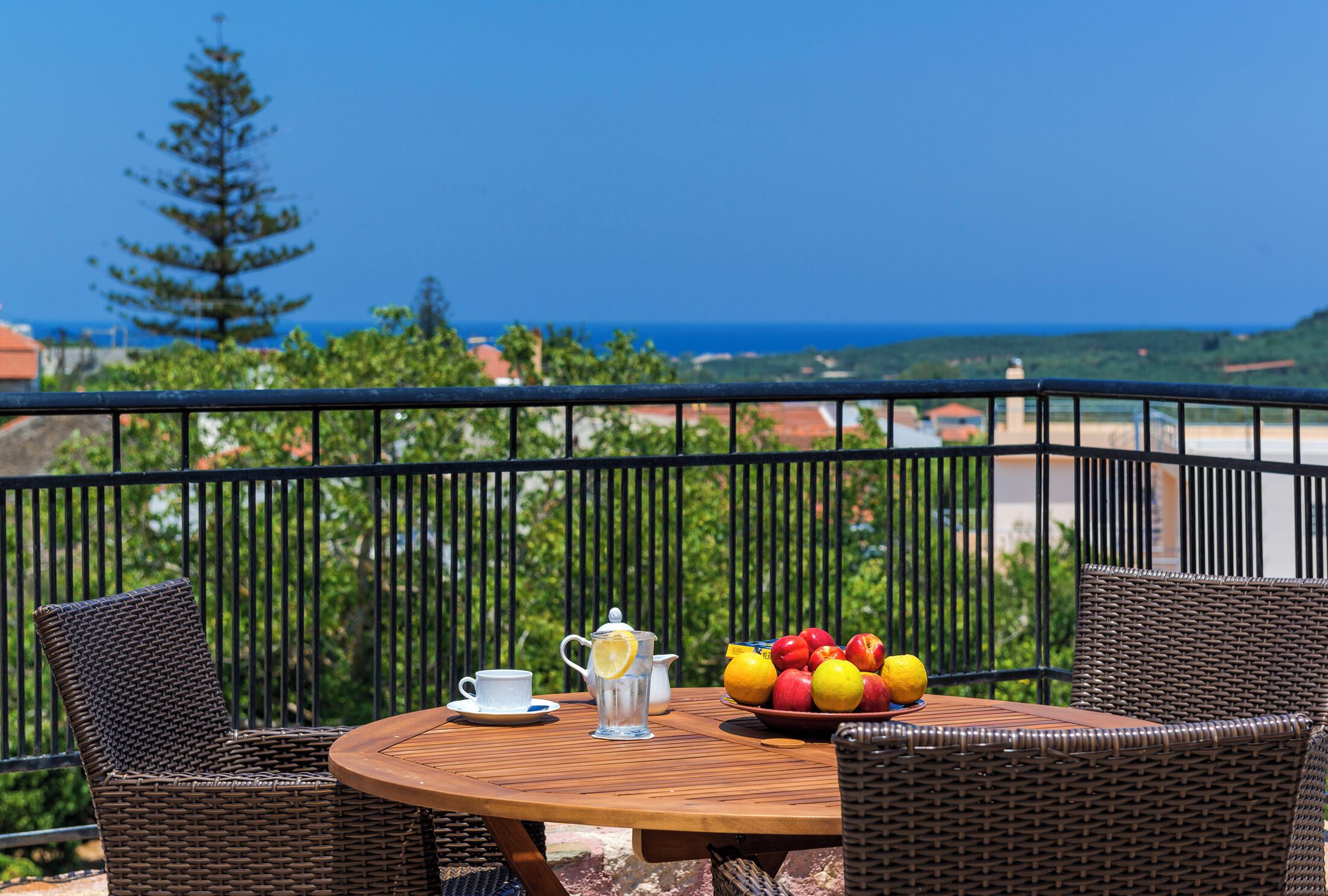 Ein runder Holztisch mit Korbstühlen, einem Wasserkrug, einer Tasse und einer Obstschale auf einem Balkon mit Blick auf das Meer und viel Grün.