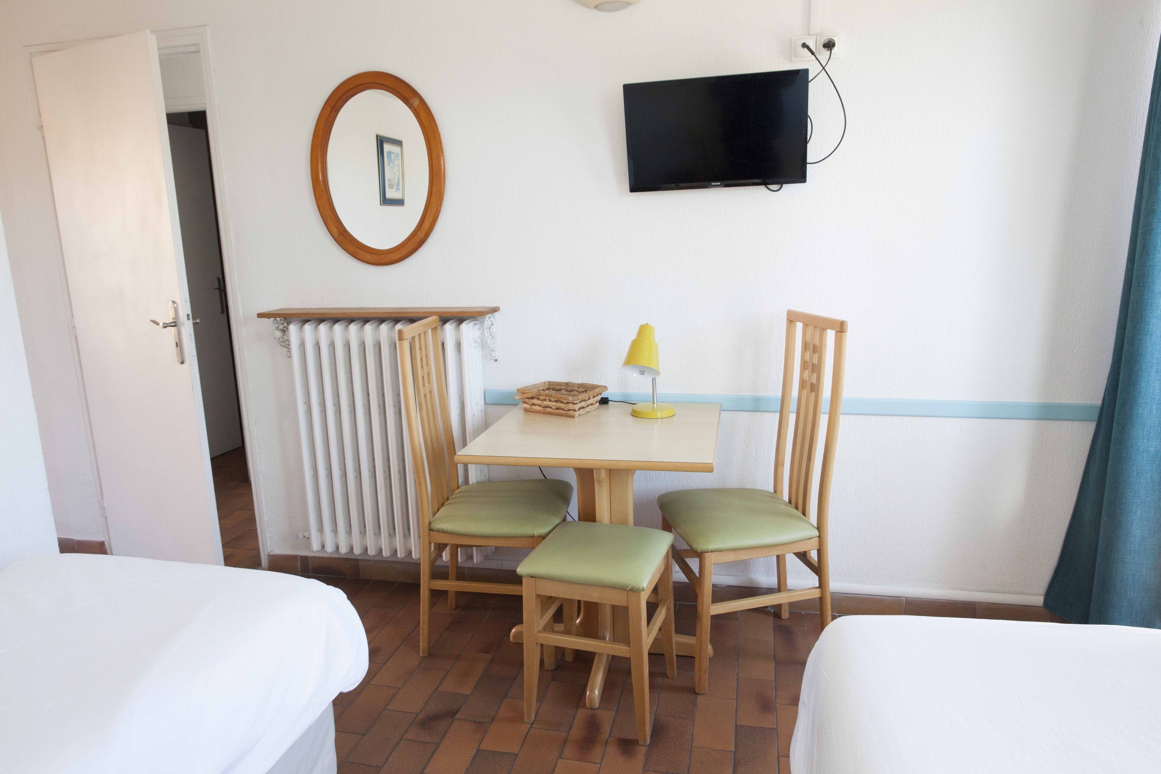 A hotel room with two beds, a table with chairs, a radiator, a TV, and a window with a curtain.