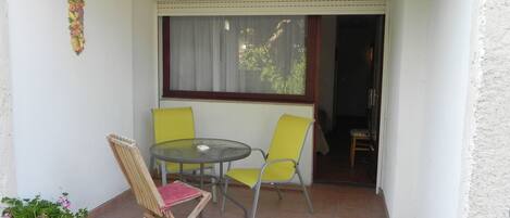 A small balcony with a table and two chairs, a potted plant, and a window with curtains.