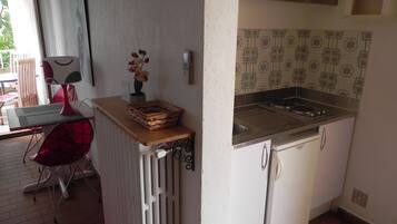 A compact kitchen with a built-in stove, a sink, and a small refrigerator. There's a chair and a table with a lamp in the adjacent dining area.