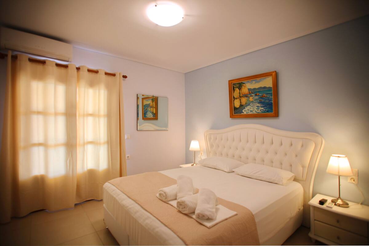 Habitación de hotel con una cama grande, una mesita de noche con lámpara, un cuadro en la pared y una ventana con cortinas.