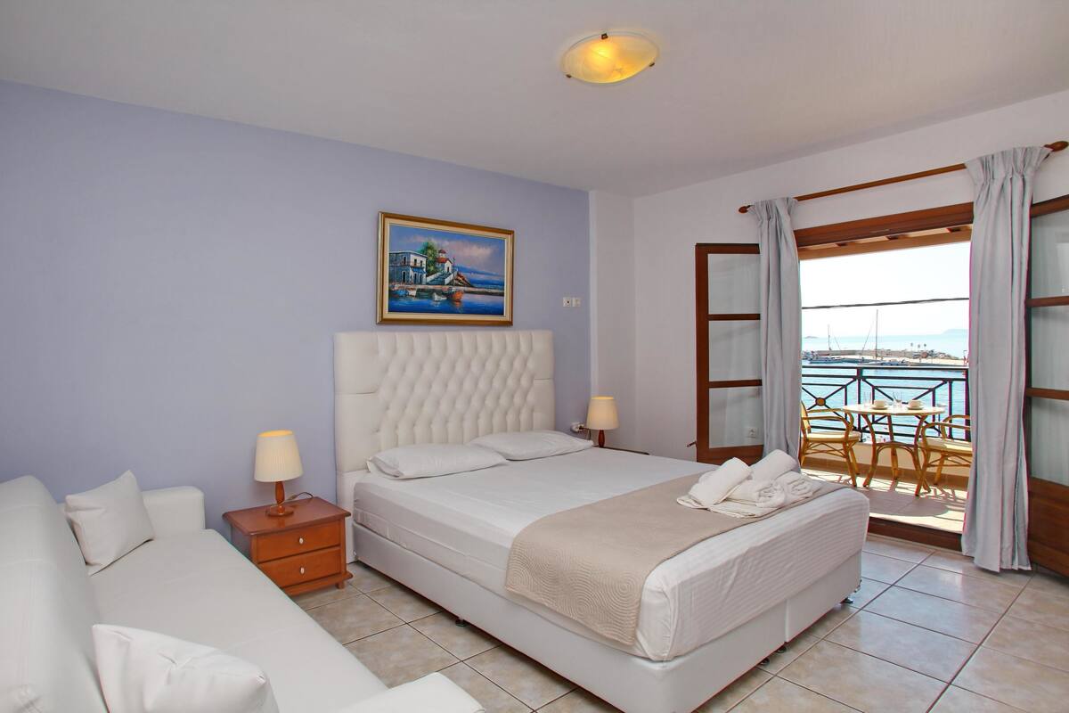 Un dormitorio con una cama grande, una mesita de noche, una lámpara, un cuadro y un balcón con vistas al mar.