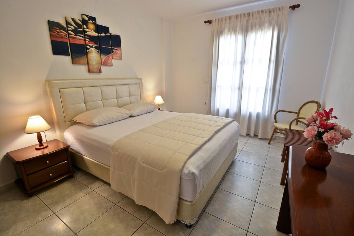 Una cama bien hecha con cabecera, una mesita de noche con lámpara, una silla y un florero con flores.