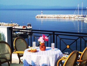 Un tavolo con tovaglia bianca, un vaso con fiori e un piatto di pasticcini su un balcone con vista su una marina, barche e un faro.