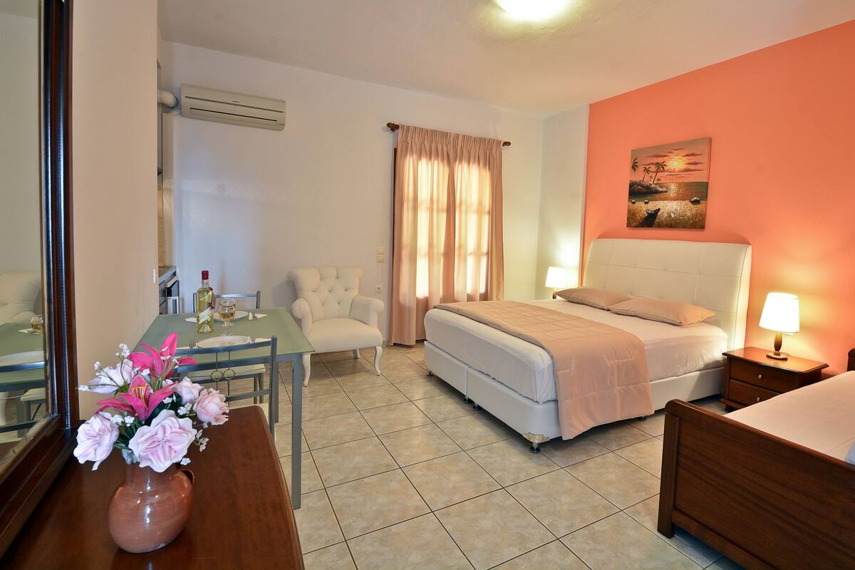 Una habitación de hotel con una cama, un escritorio con una silla, una mesita de noche con una lámpara, un espejo y un jarrón con flores.