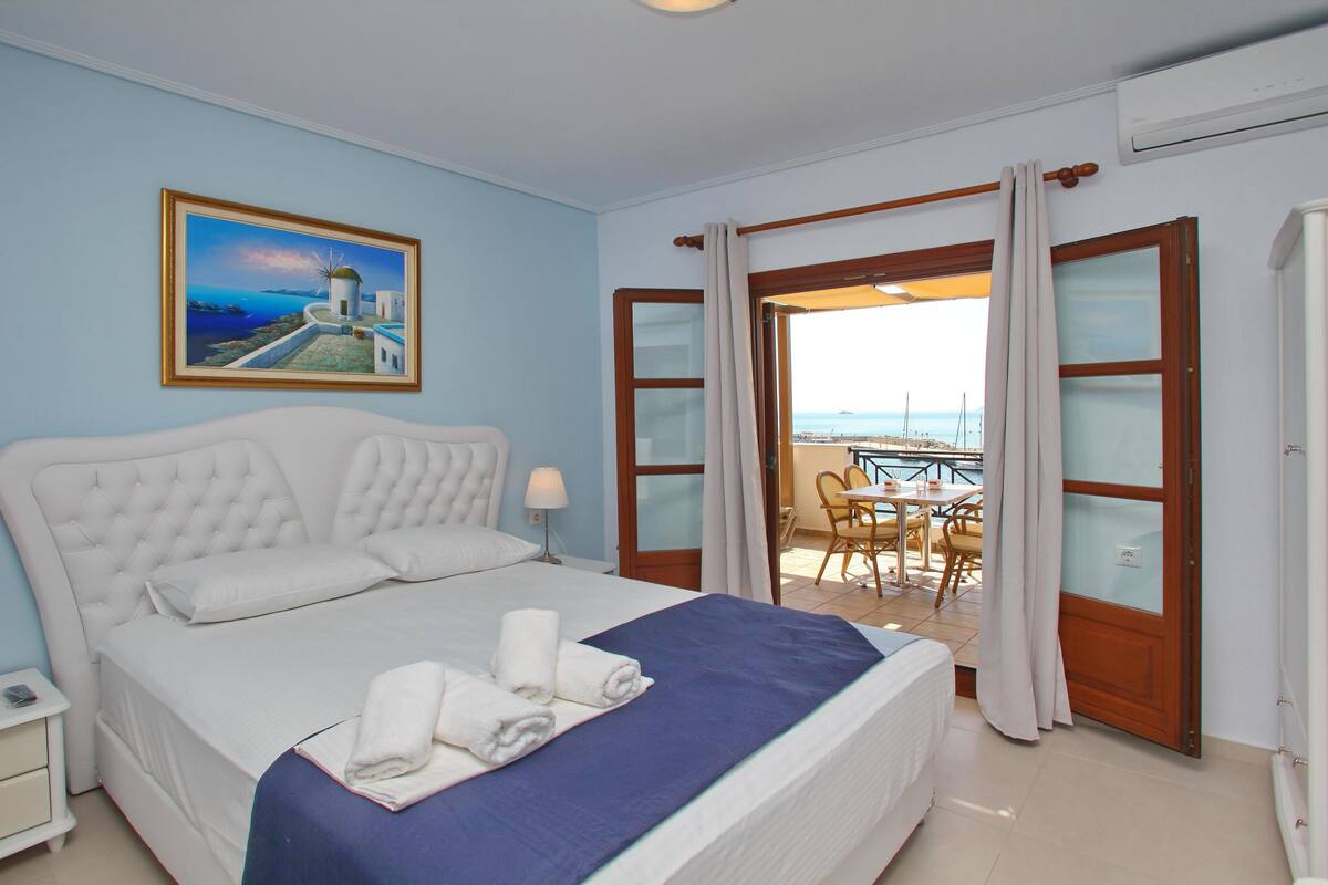 Un dormitorio con cama, mesita de noche, un cuadro de una escena costera y vistas al mar a través de una puerta abierta.