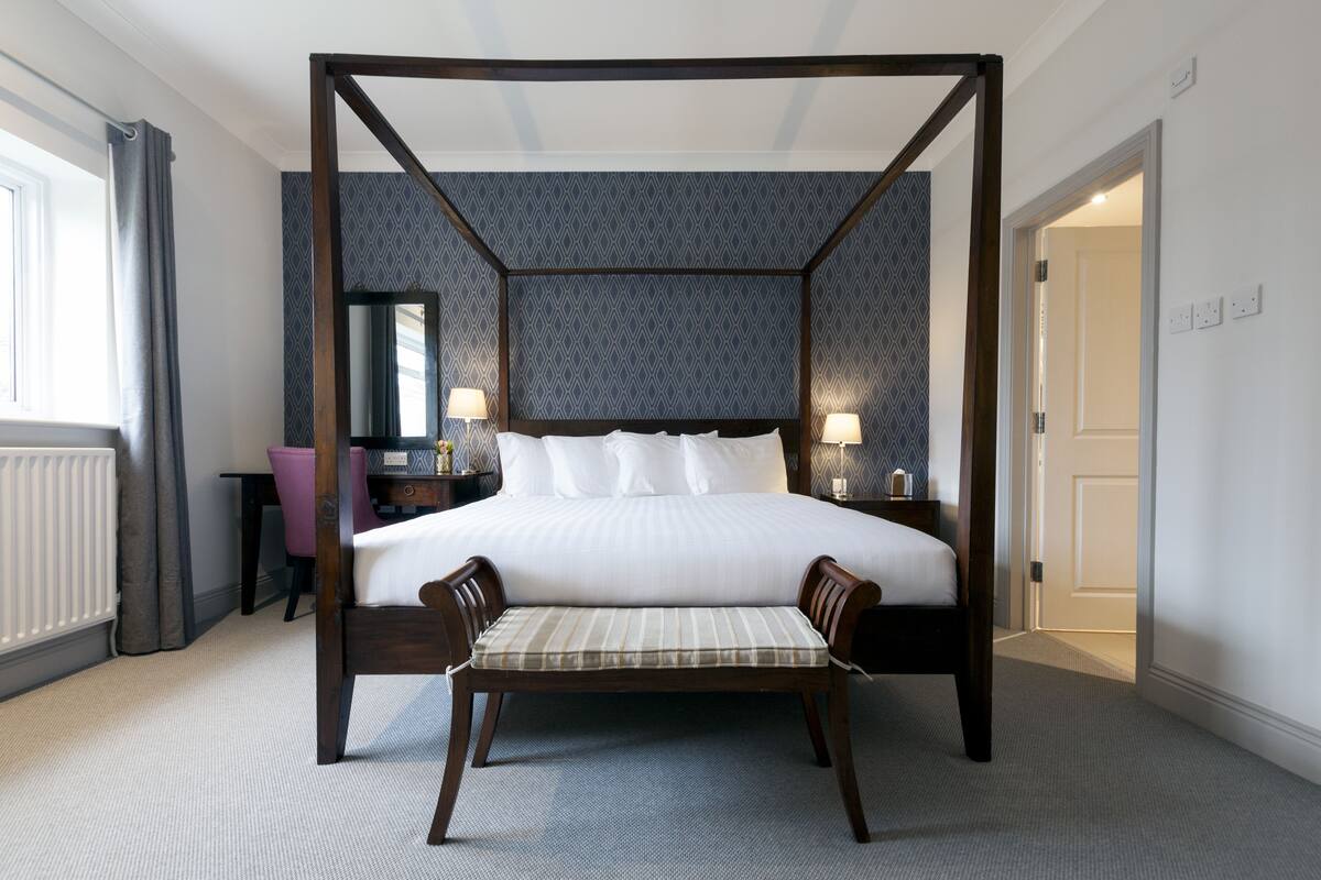 A four-poster bed with a headboard, a bedside table with a lamp, a chair with a cushion, and a mirror on the wall.