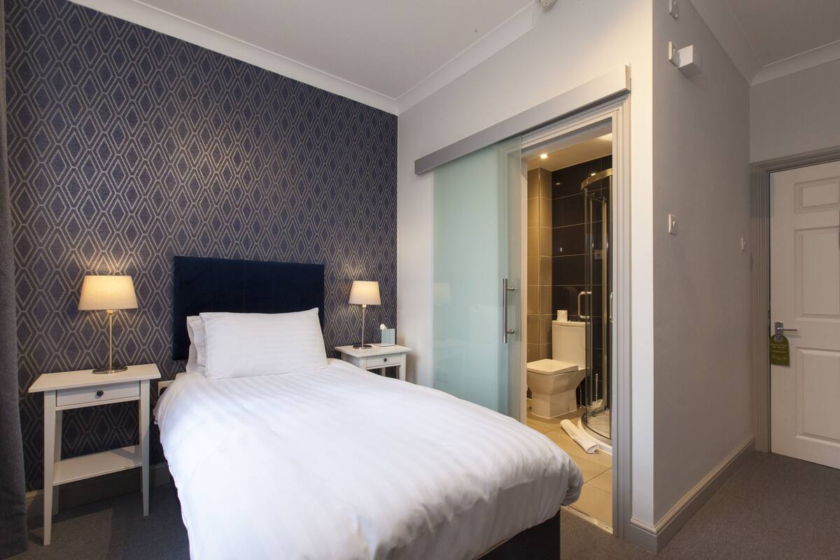 A hotel room with a bed, two bedside tables with lamps, a shower area, and a door leading to another room.