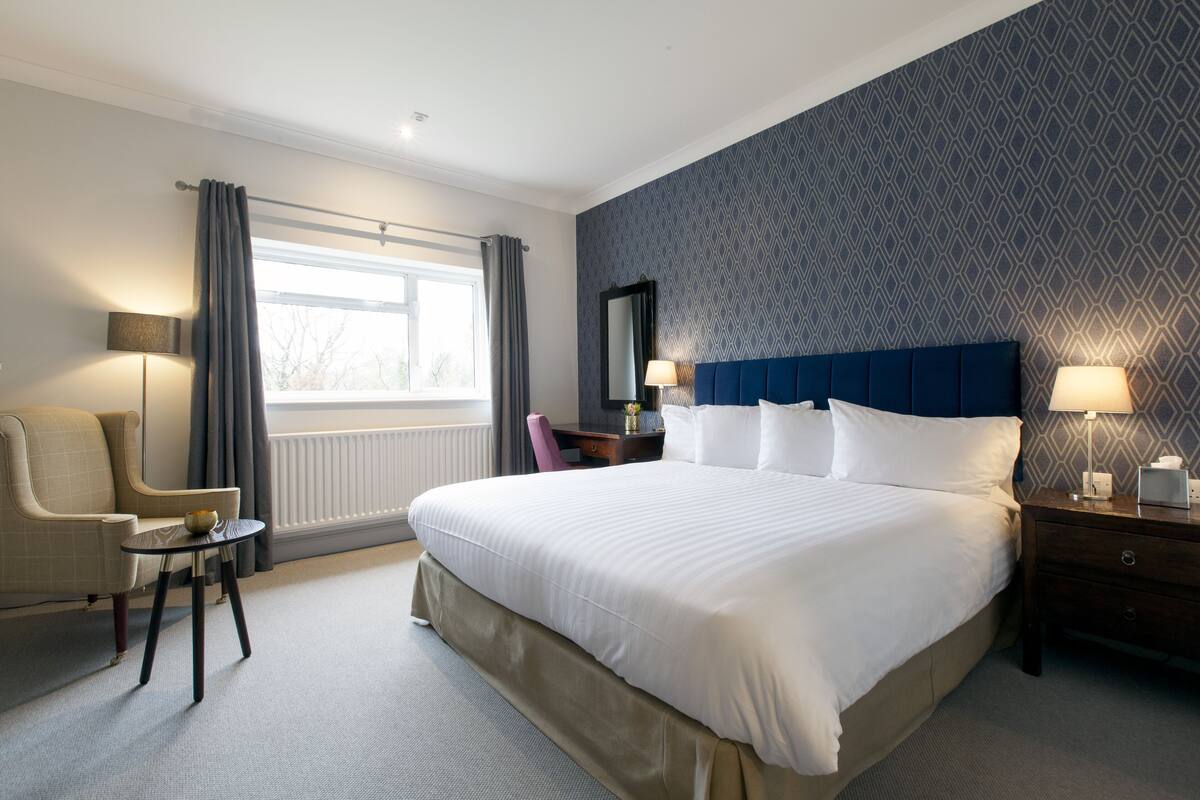 A hotel room with a large bed, a chair, a small table, a nightstand, and a window with curtains.