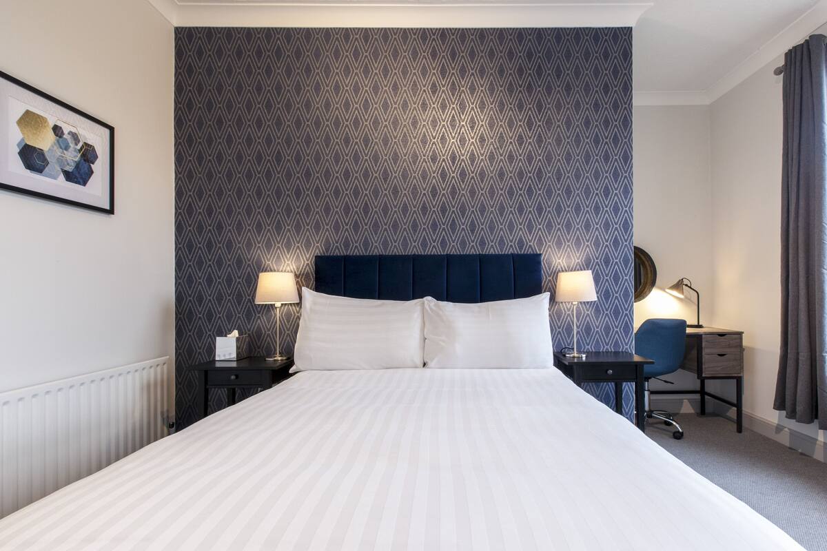 A hotel room with a large bed, two bedside tables with lamps, a desk with a chair, and a patterned wallpaper.