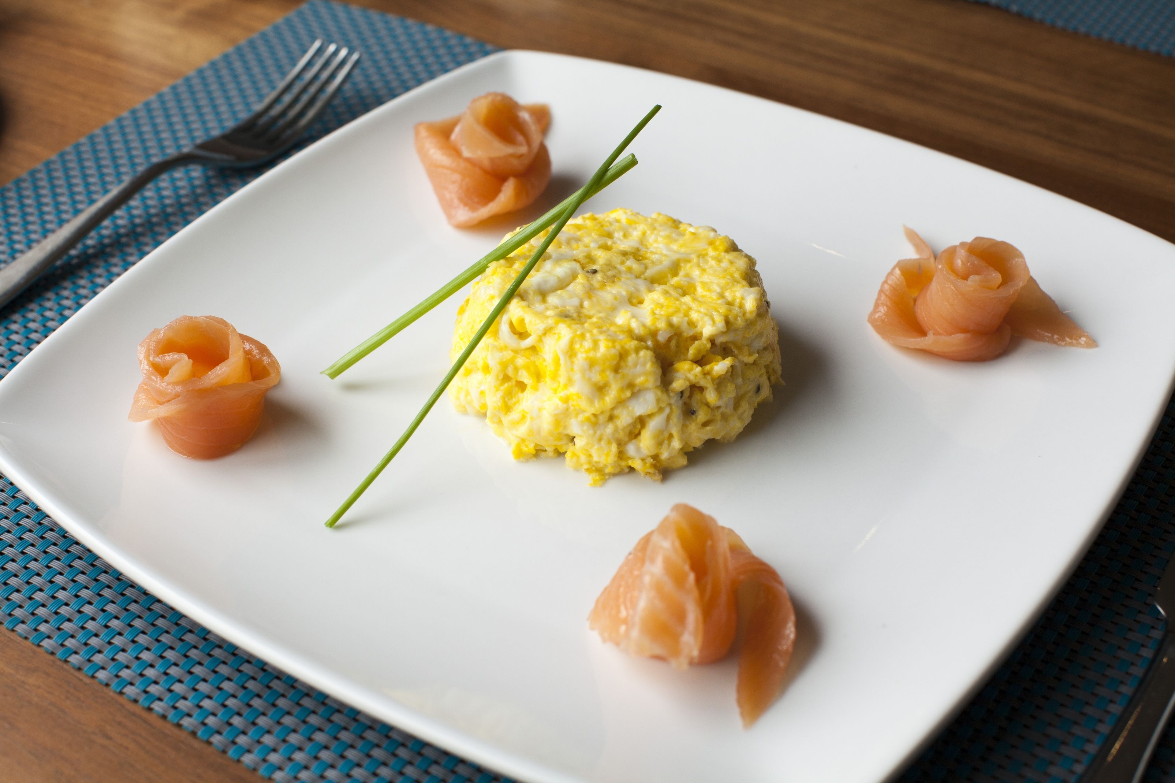 Un plato blanco con huevos revueltos, salmón ahumado y cebollín sobre un camino de mesa azul.