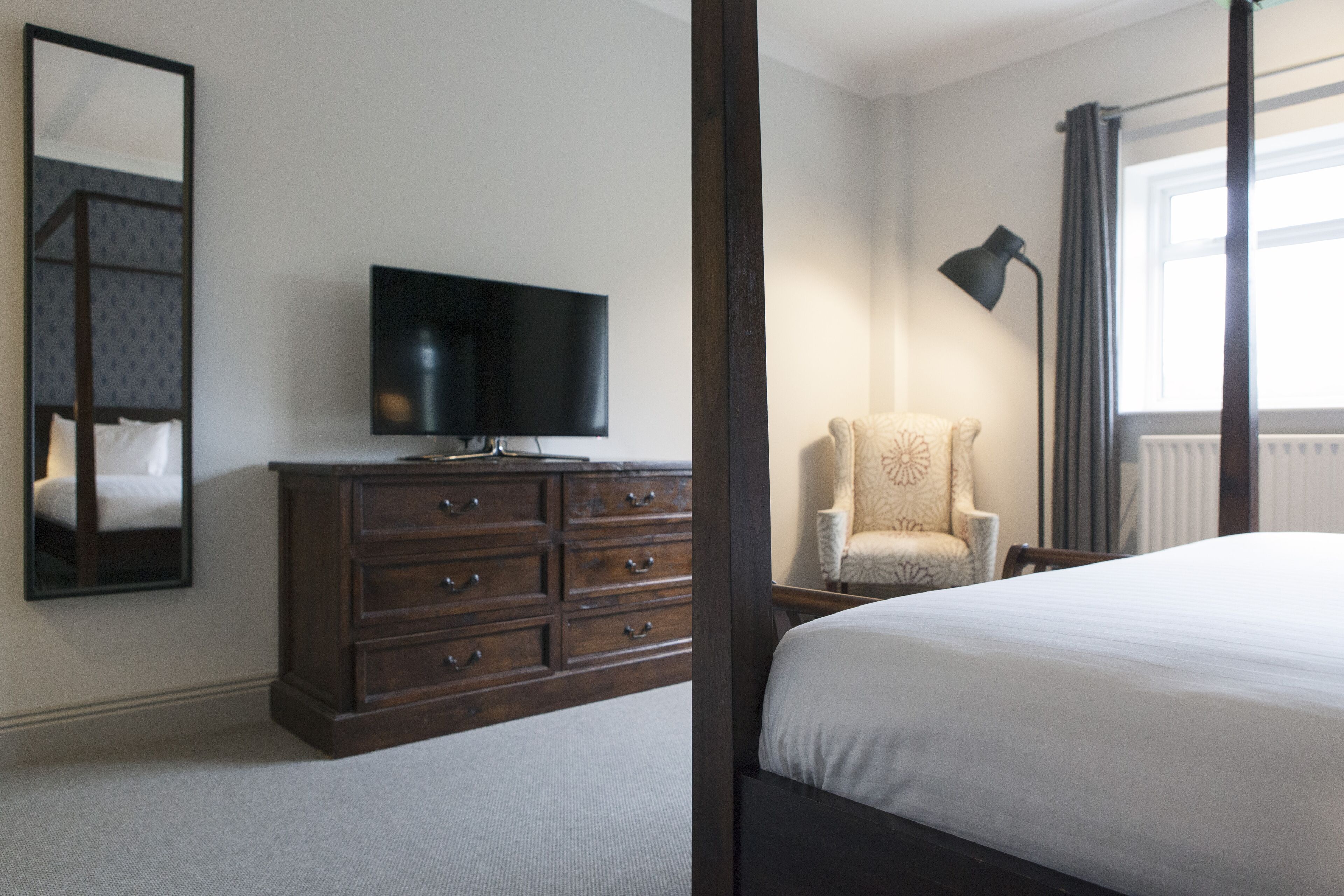 Une chambre à coucher avec un lit à baldaquin, un téléviseur posé sur une commode, une chaise et une fenêtre avec des rideaux.
