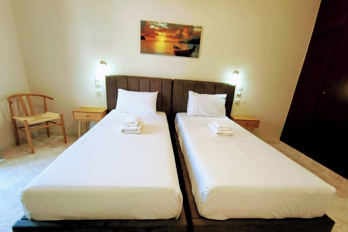 A double bed with white linens, a wooden chair, a small table, and a painting on the wall.