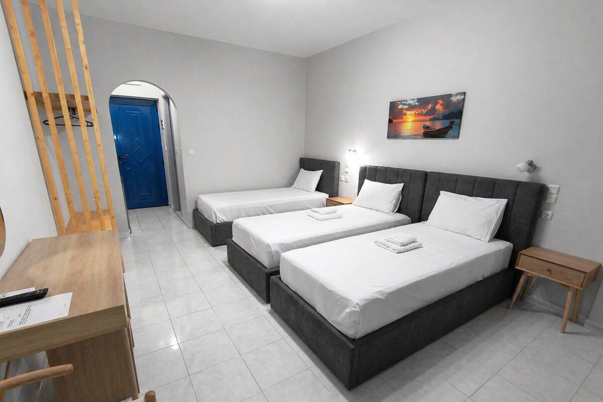 A hotel room with two beds, a desk, a chair, a wooden bench, a blue door, and a sunset painting on the wall.