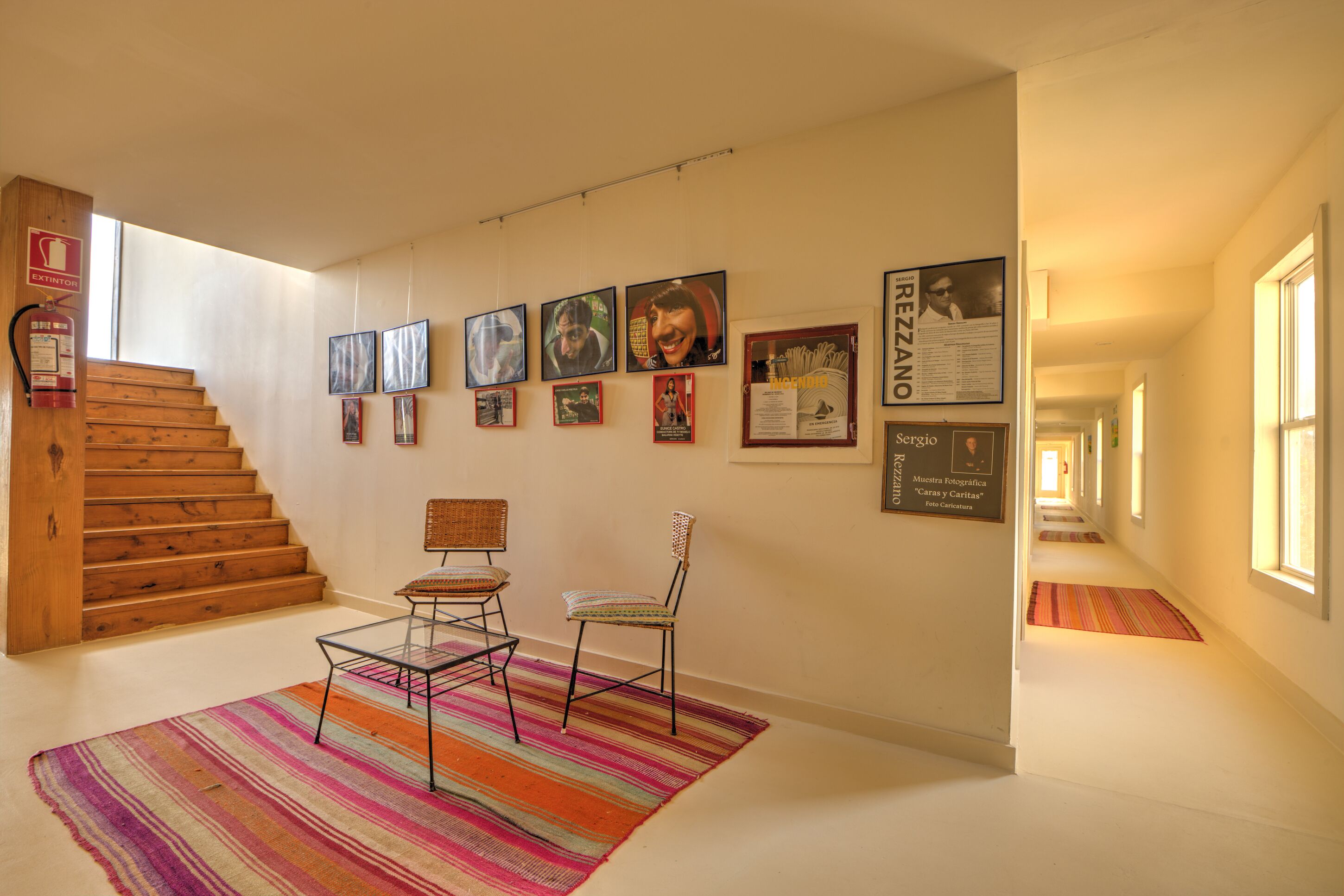 A hallway with a staircase, framed pictures on the wall, and two chairs with a colorful rug.