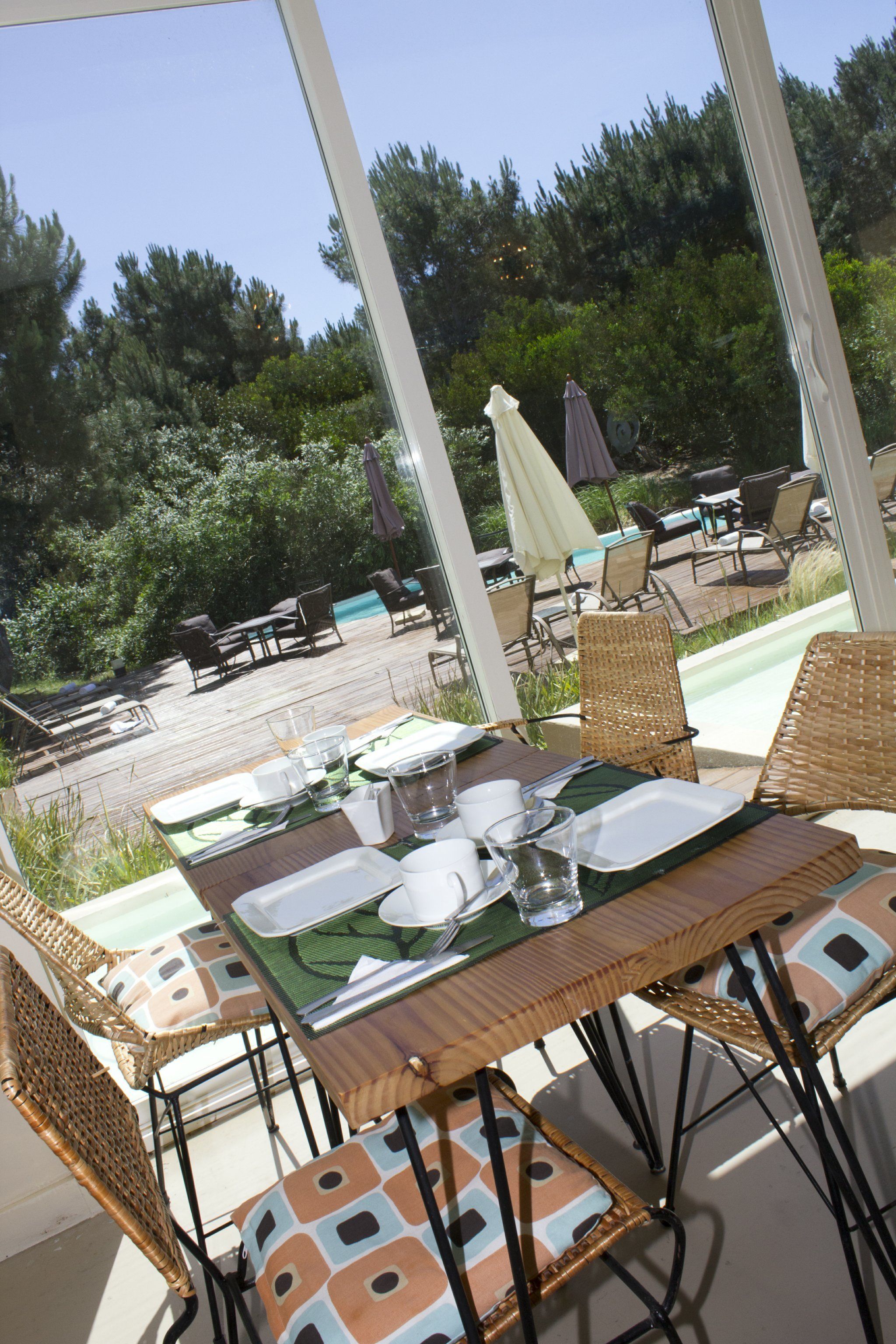 Un área de comedor al aire libre con mobiliario de mimbre, una mesa preparada con platos, vasos y cubiertos, y vistas a una piscina y follaje al fondo.