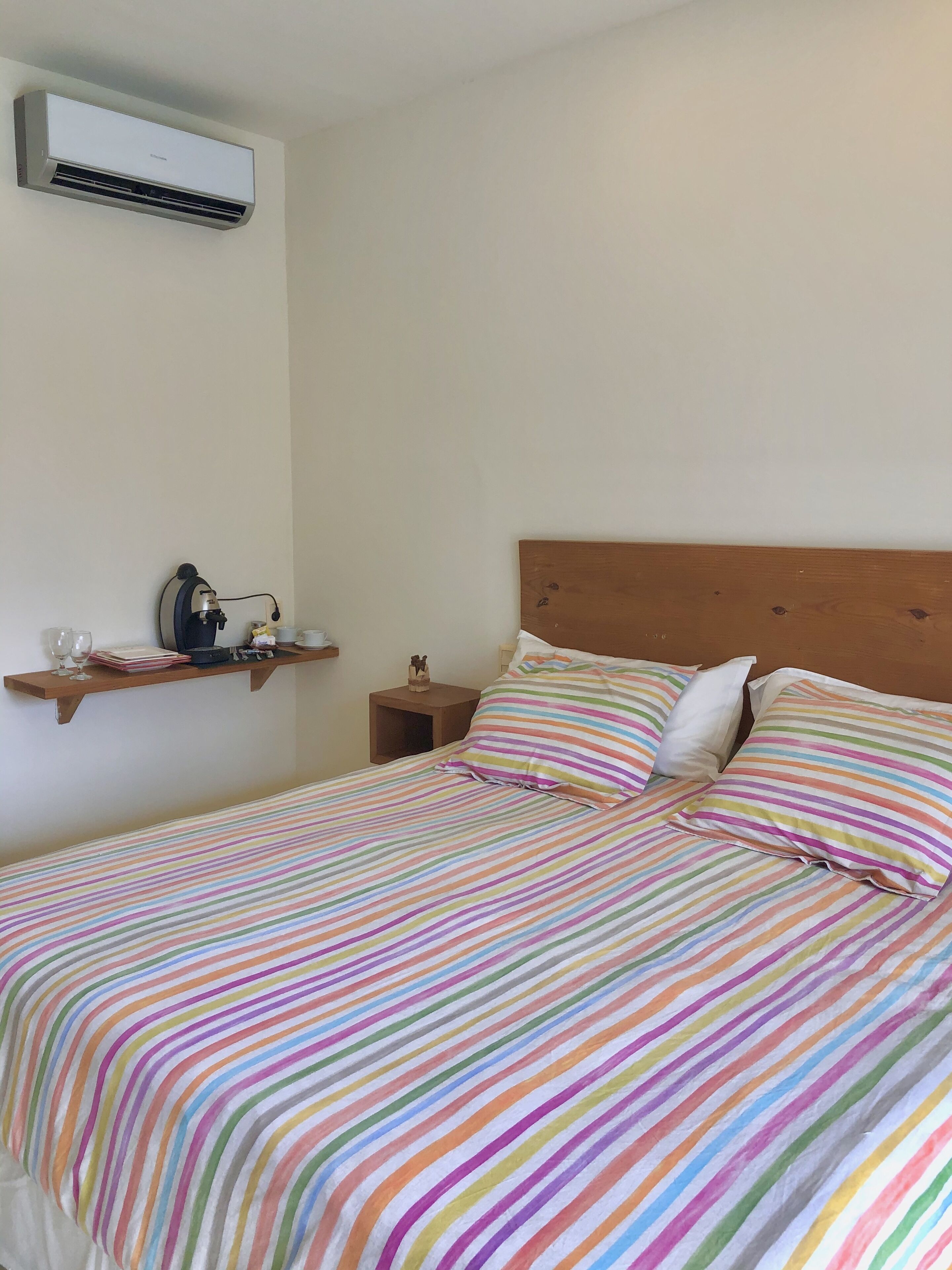 Une chambre à coucher avec un lit, une literie à rayures colorées, une tête de lit en bois, une table de chevet, une étagère avec divers objets et un appareil de climatisation.