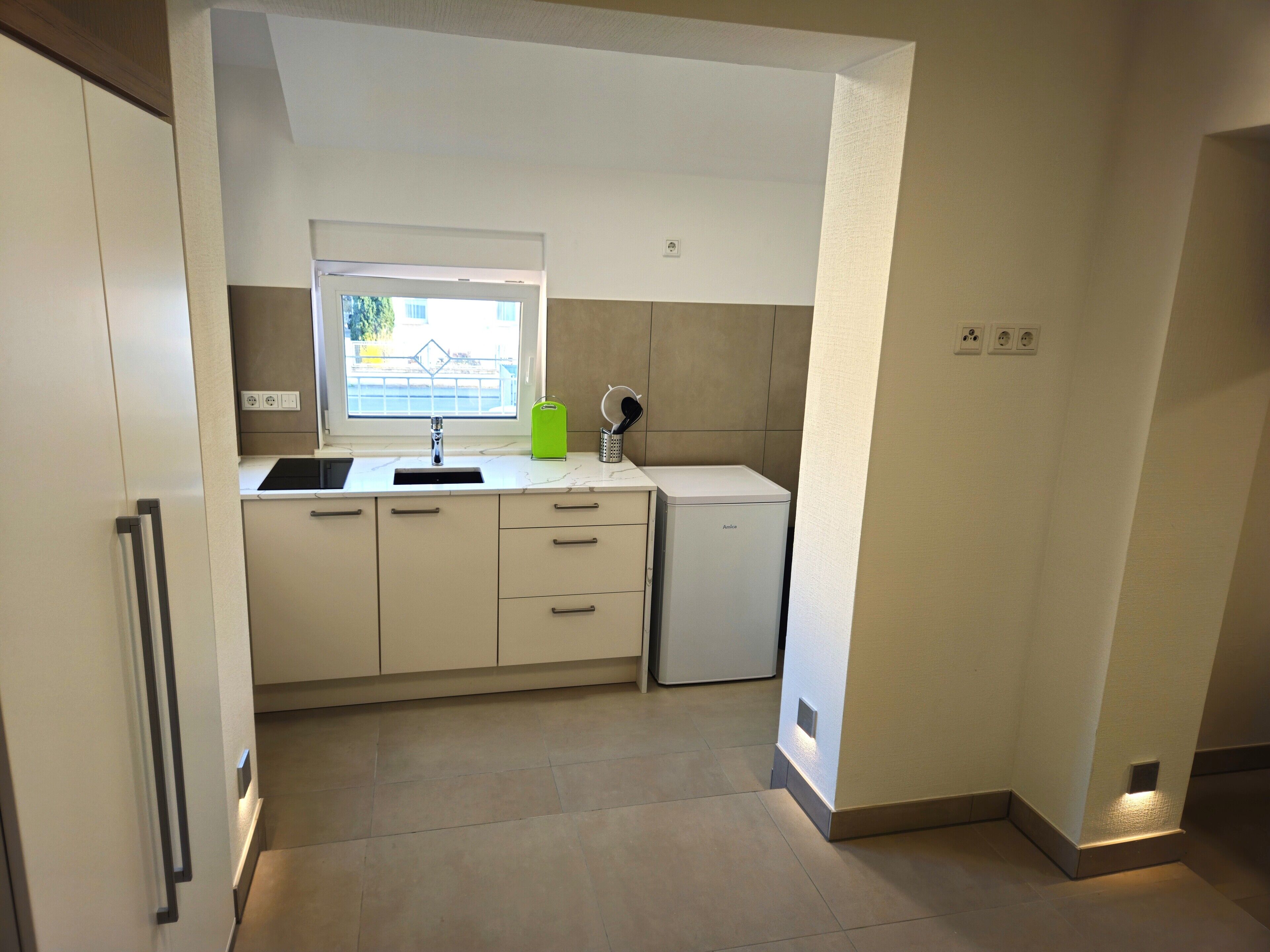 A compact kitchen with a window, a sink, a small refrigerator, and a countertop with a green container.