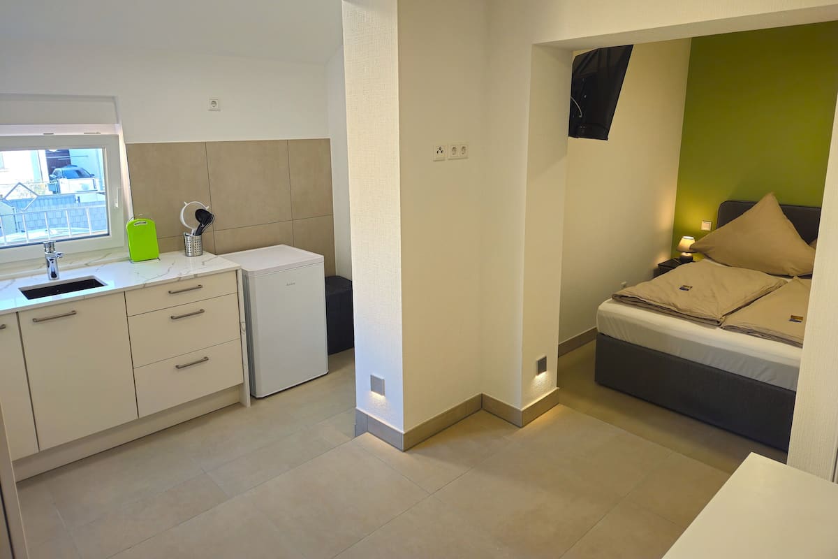 Ett kompakt hotellrum med ett litet pentry, en säng med beige sängkläder och en grön accentvägg.