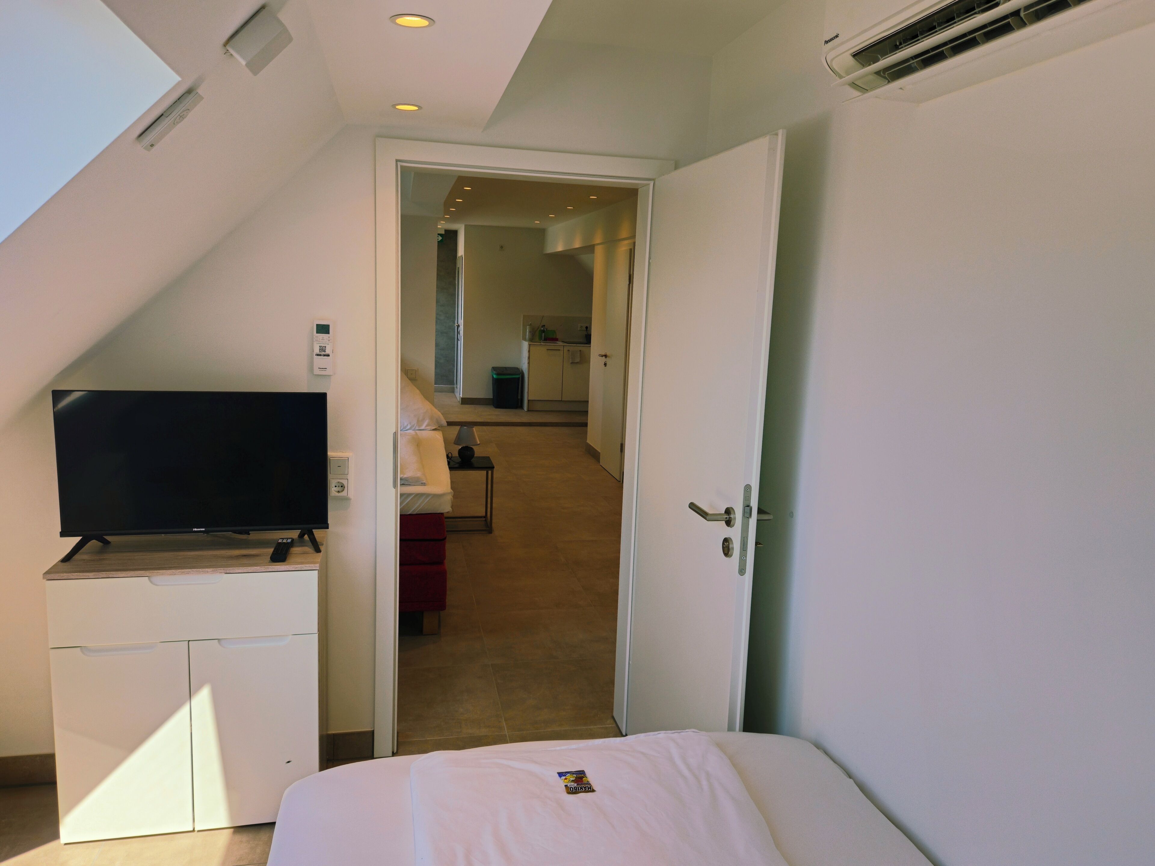 Ein Hotelzimmer mit Fernseher auf einem Tisch, einem Bett, einer Tür zu einem Flur und einem Deckenlüfter.