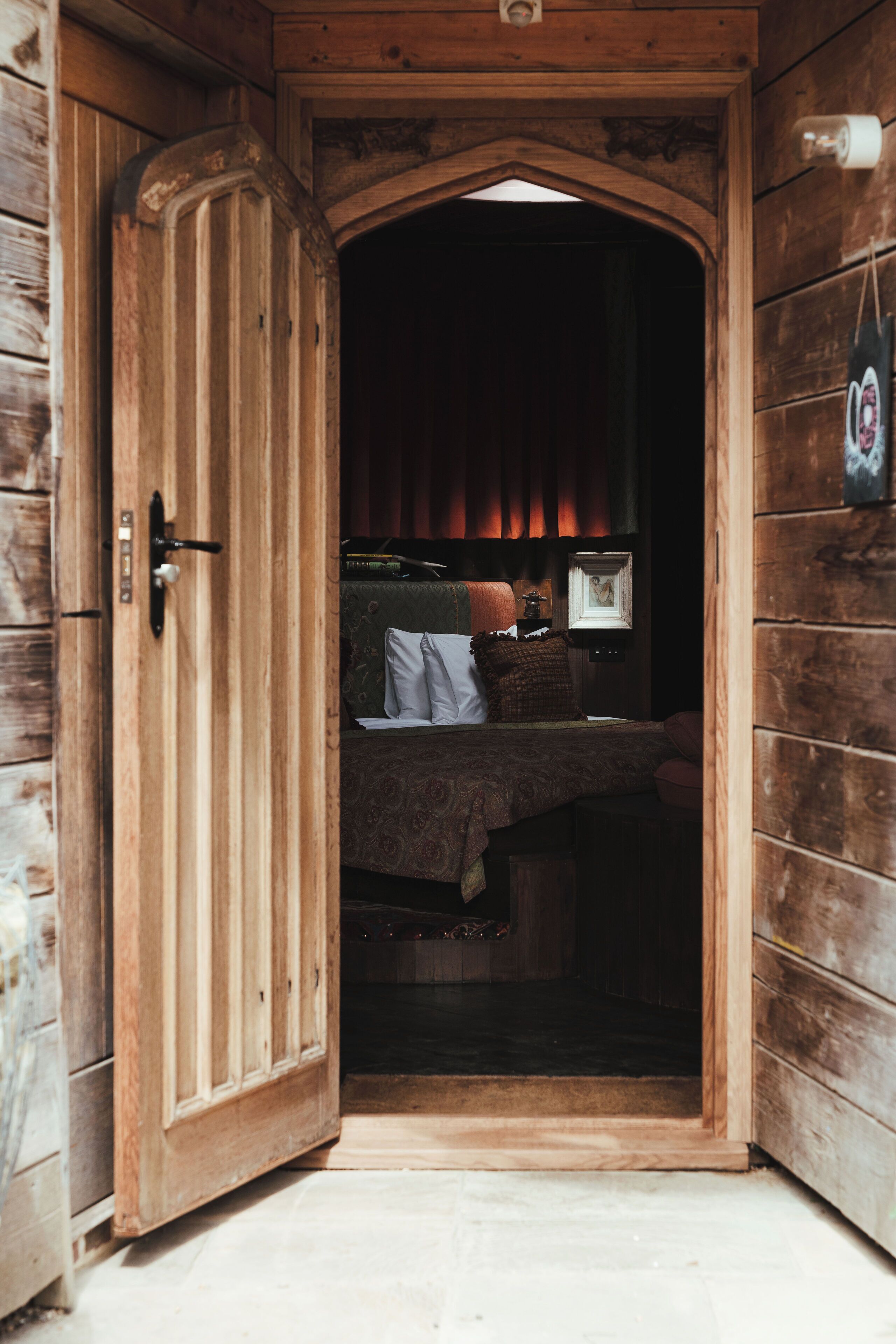 Uma porta de madeira que dá para um quarto com uma cama, um criado-mudo e uma luminária na parede.