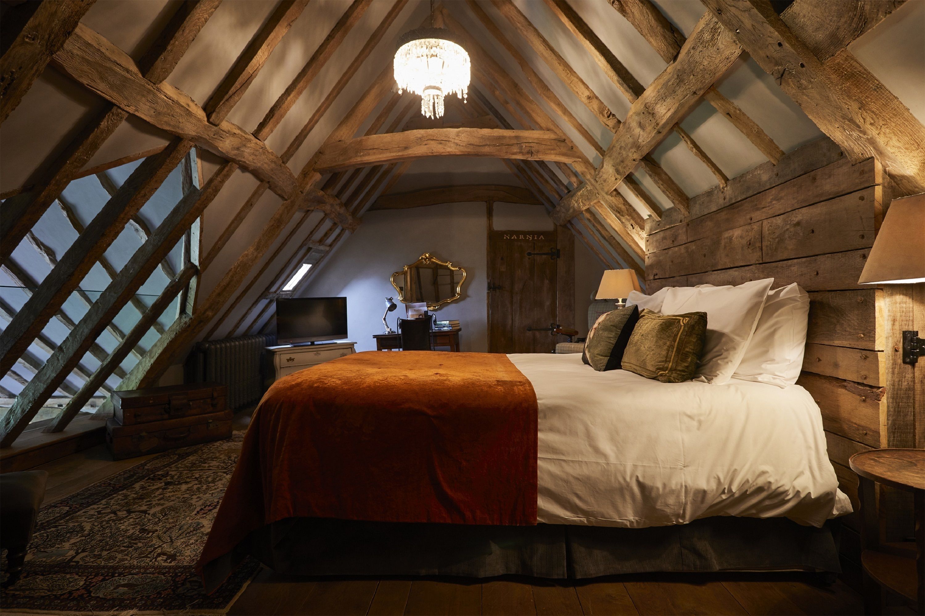 A bedroom with a sloped ceiling, a chandelier, a bed with white linens, a mirror, and a television.