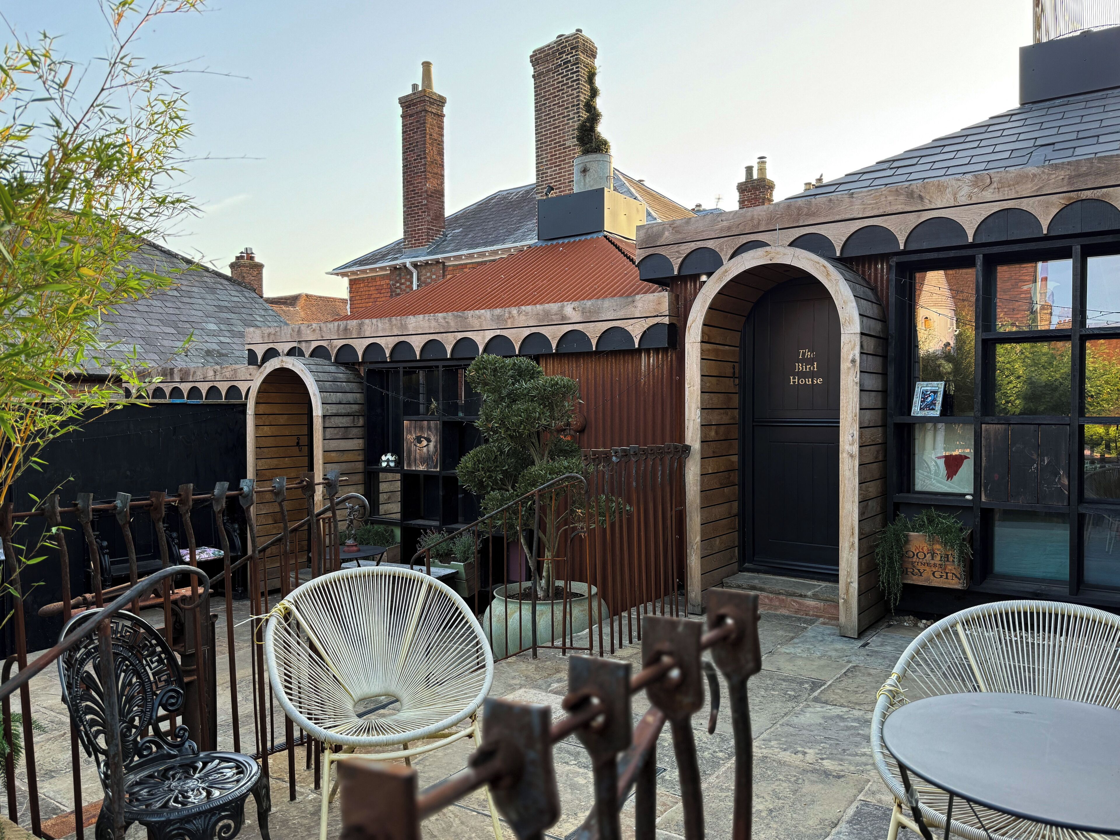Un patio con sillas de mimbre y una mesa, un edificio con un cartel que dice "The Bird House", y chimeneas de ladrillo.