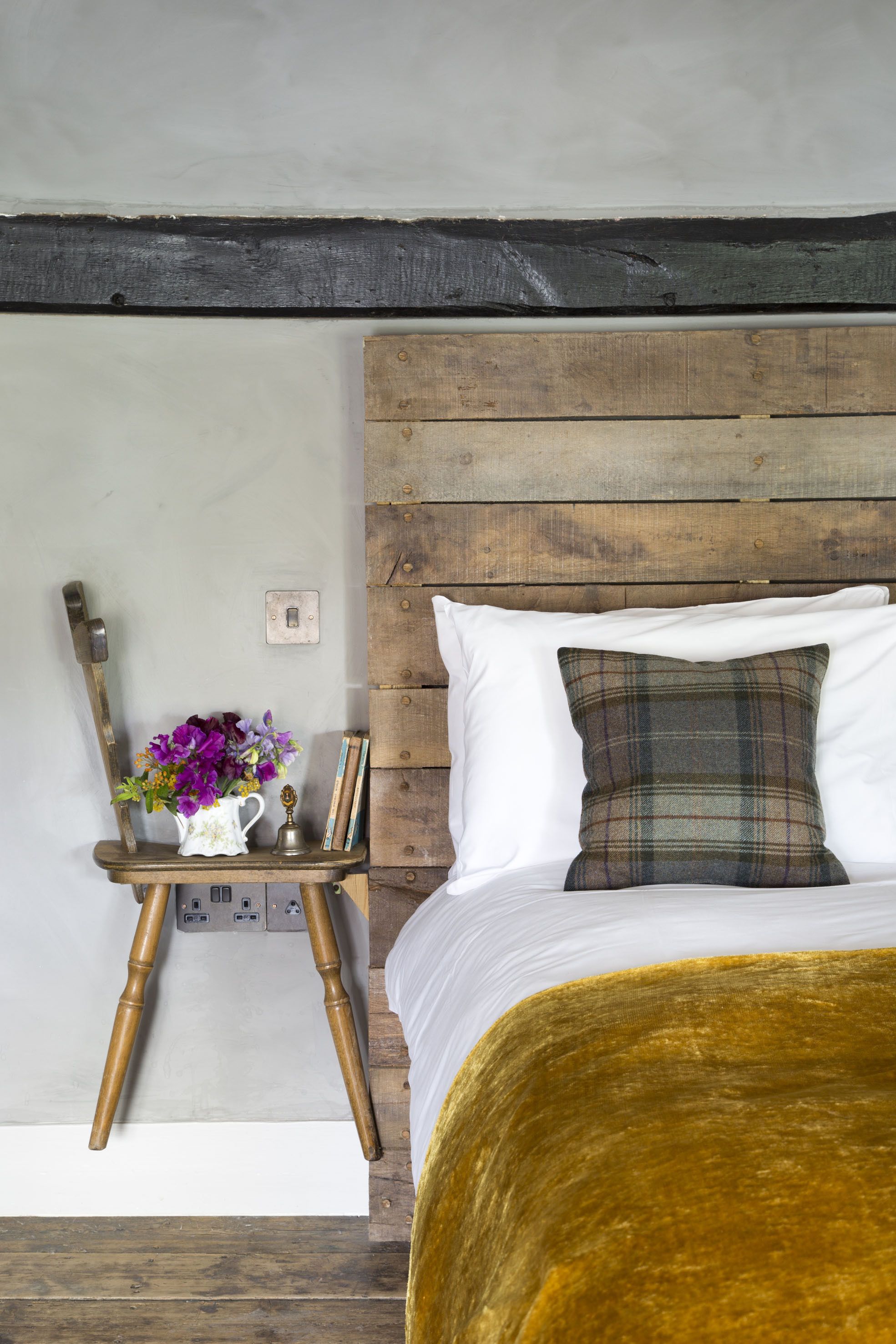A bedroom with a wooden headboard, a bed with a yellow blanket, a nightstand with a vase of flowers, and a lamp.