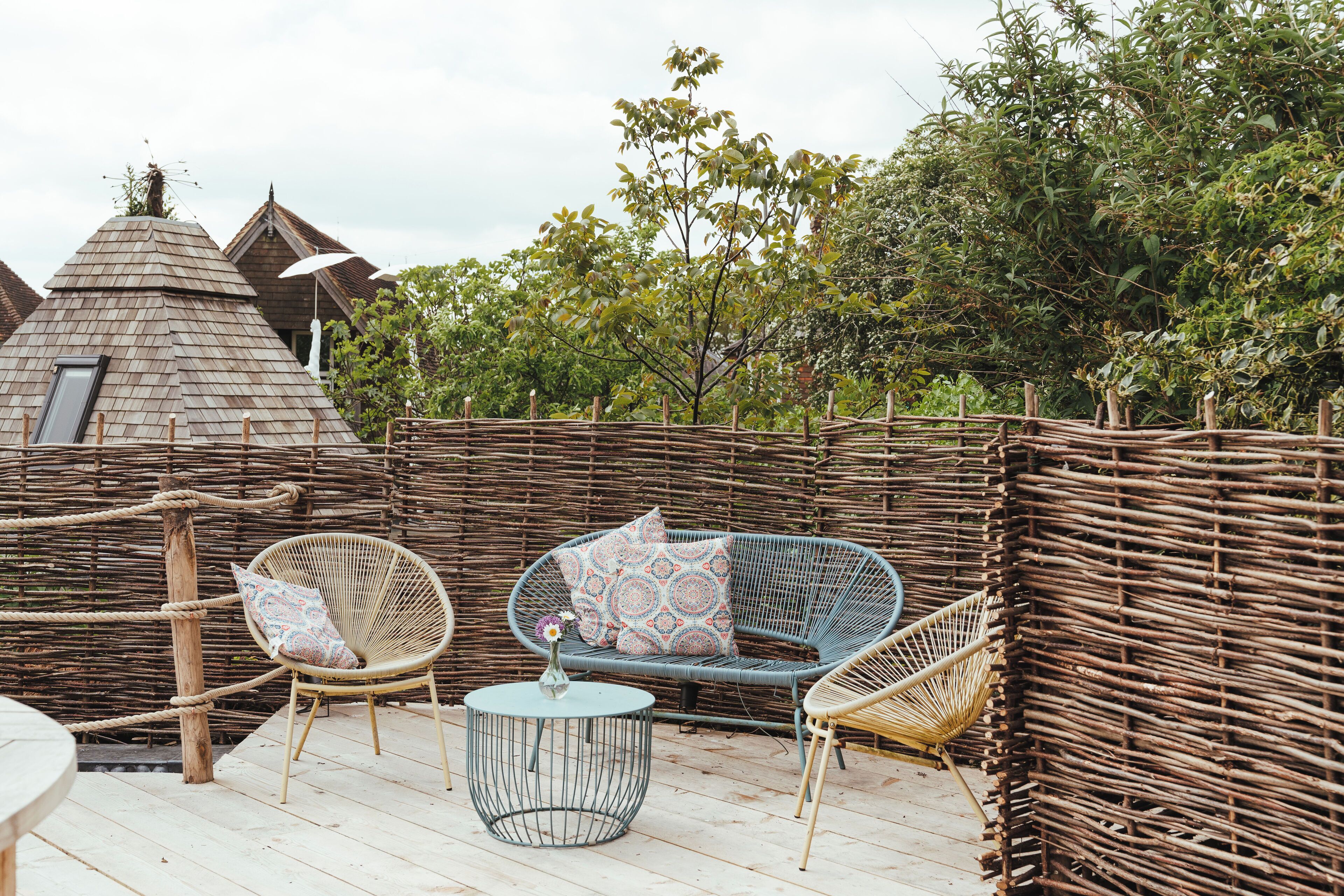 Een houten terras met rieten meubels, waaronder een bank en stoelen, en een kleine ronde tafel met een vaas.