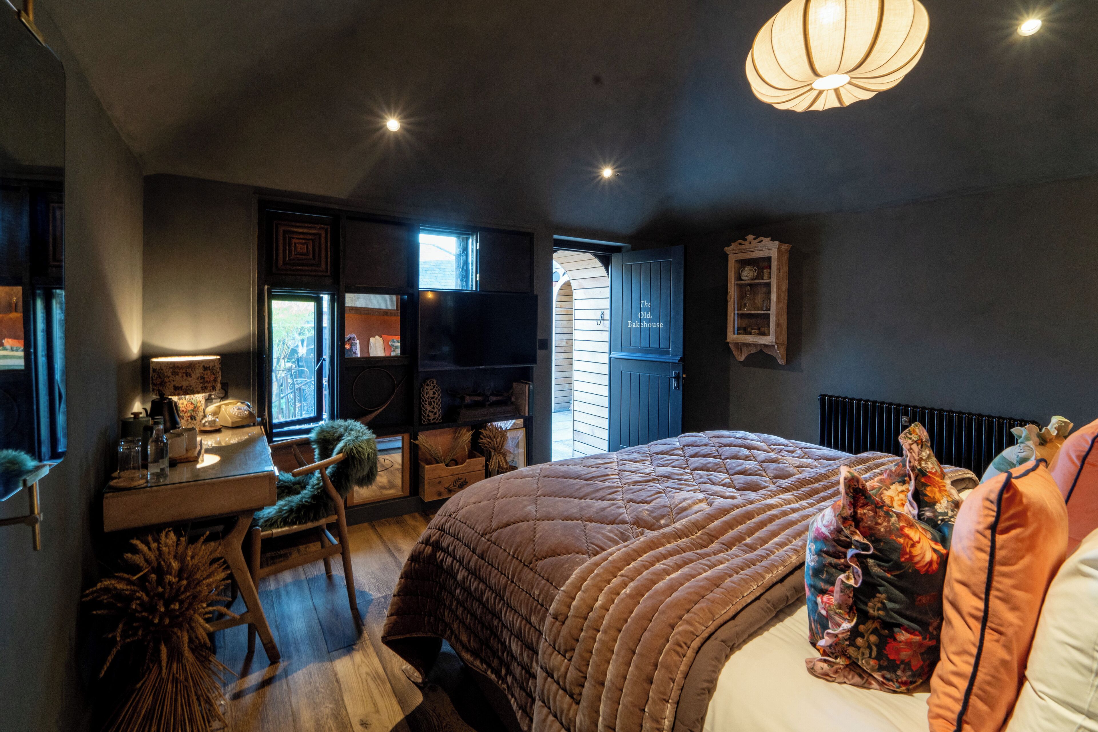 Une chambre avec un grand lit, un bureau et une chaise. Il y a une fenêtre et une porte qui donne sur une autre pièce.