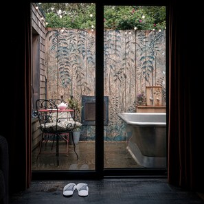 A room with a sliding glass door leading to a garden. Inside, there is a chair with a cushion, a small table with a teapot, and a bathtub.