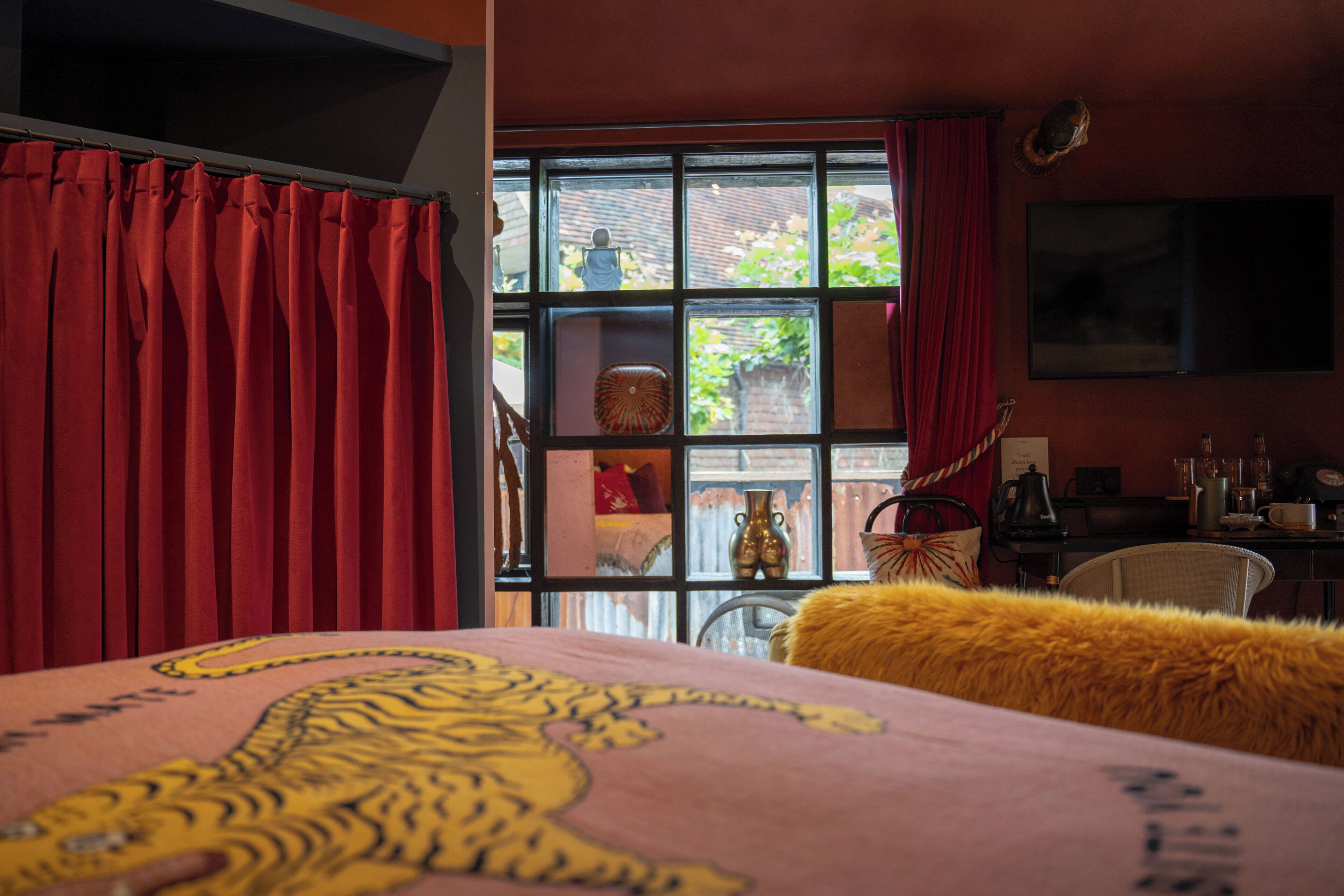 A room with a bed featuring a tiger design, a window with red curtains, and a view of the outside.