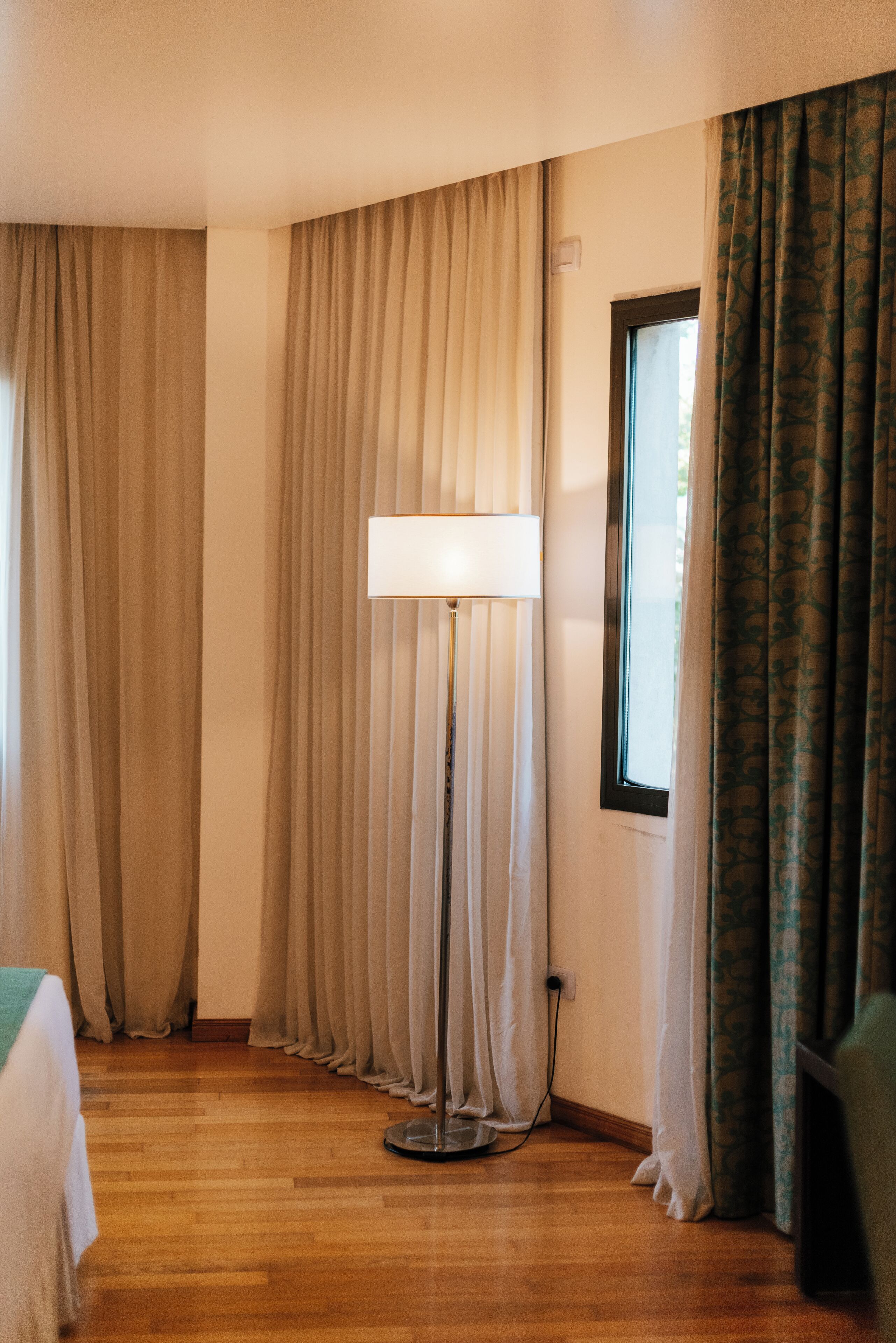 A hotel room with a wooden floor, a standing floor lamp, beige curtains, and a window with patterned curtains.