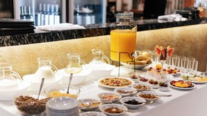 Tarjolla on runsas buffetpöytä, jossa on monenlaista ruokaa ja juomaa, kuten suuri kannu appelsiinimehua, kulhoja täynnä muroja ja lautasellisia hedelmiä.