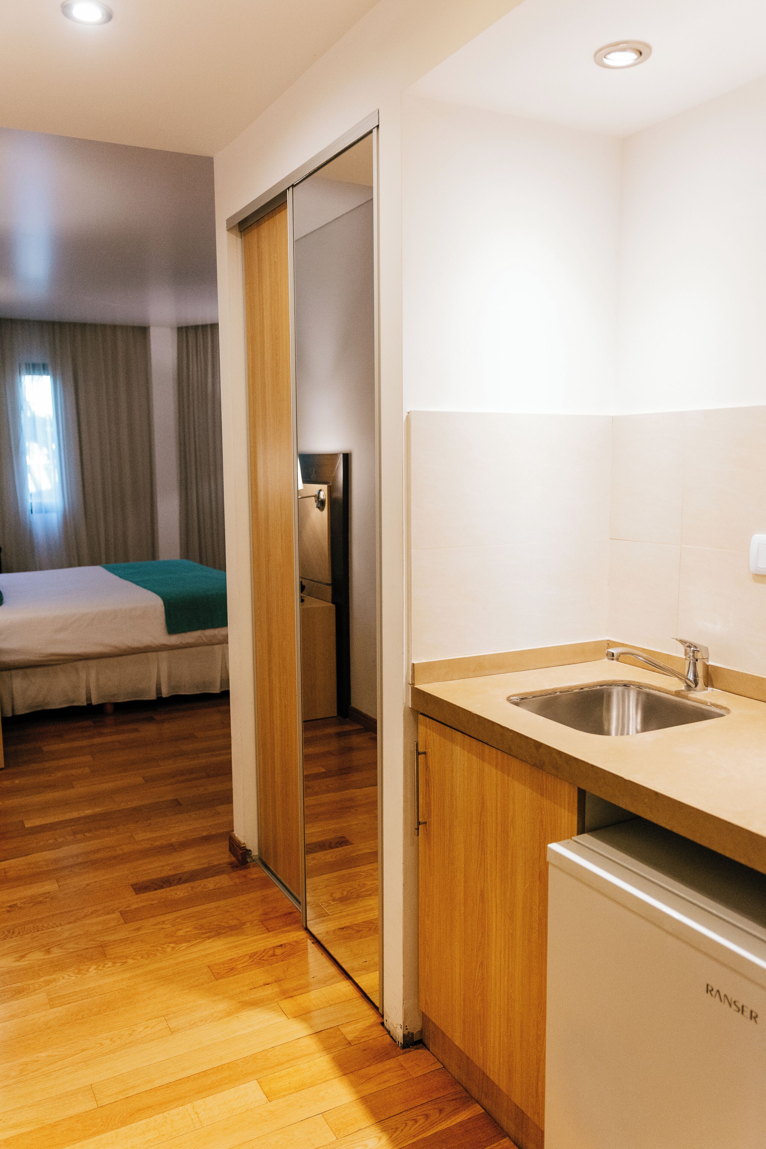 A compact hotel room with a wooden floor, a bed with a green blanket, a kitchenette with a sink, and a refrigerator.