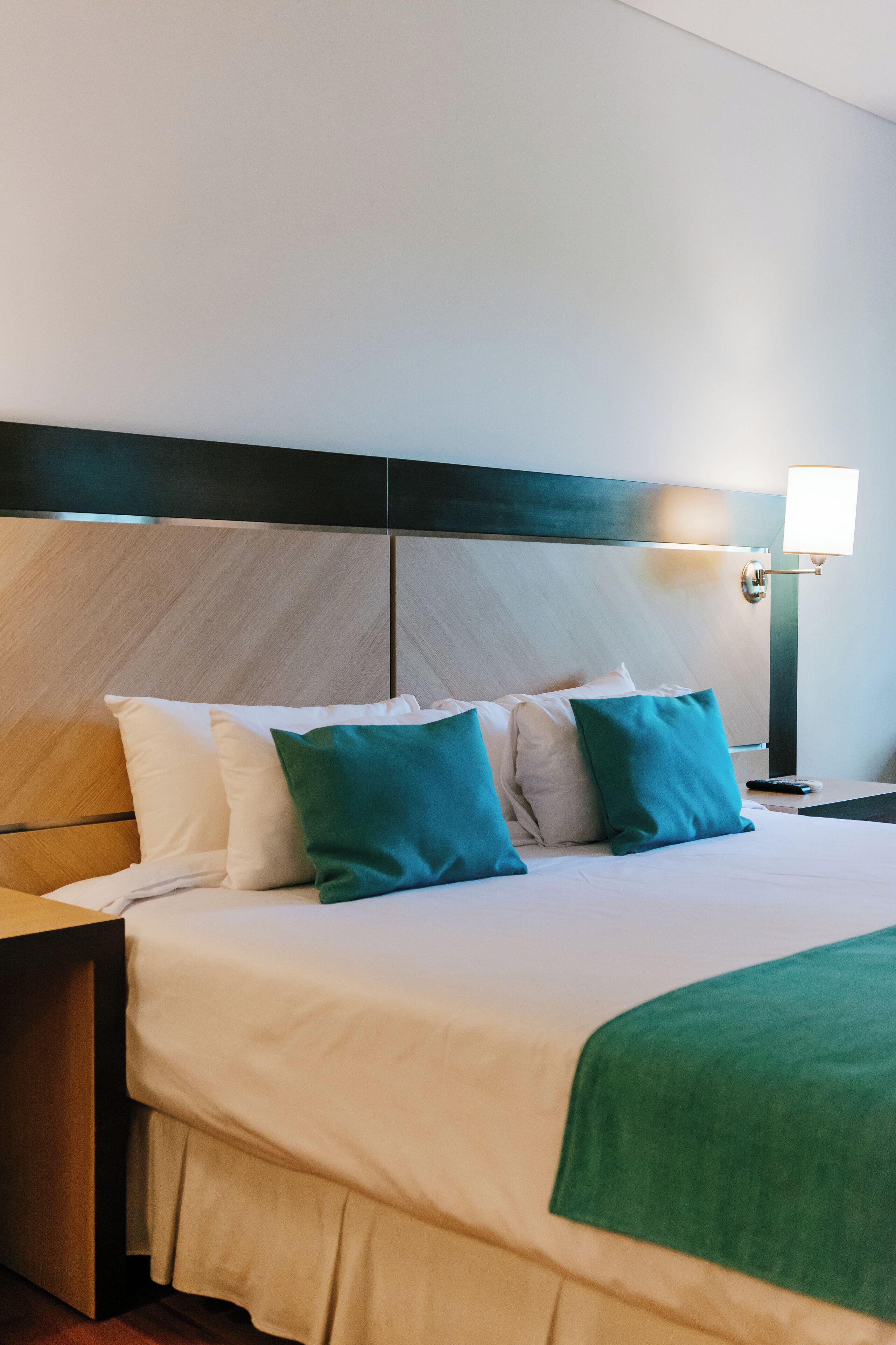 A neatly made bed with white and green pillows, a wooden headboard, and a bedside lamp.