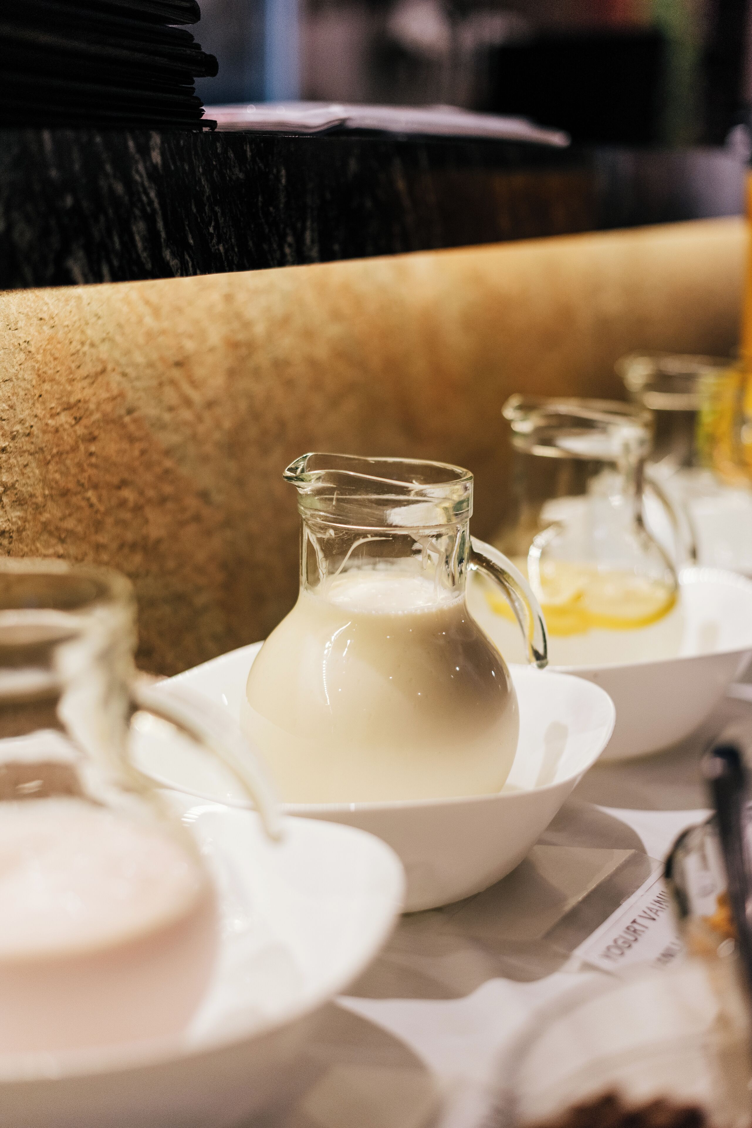 A close-up of a table with a pitcher of milk, a bowl, and a glass pitcher on a tablecloth.