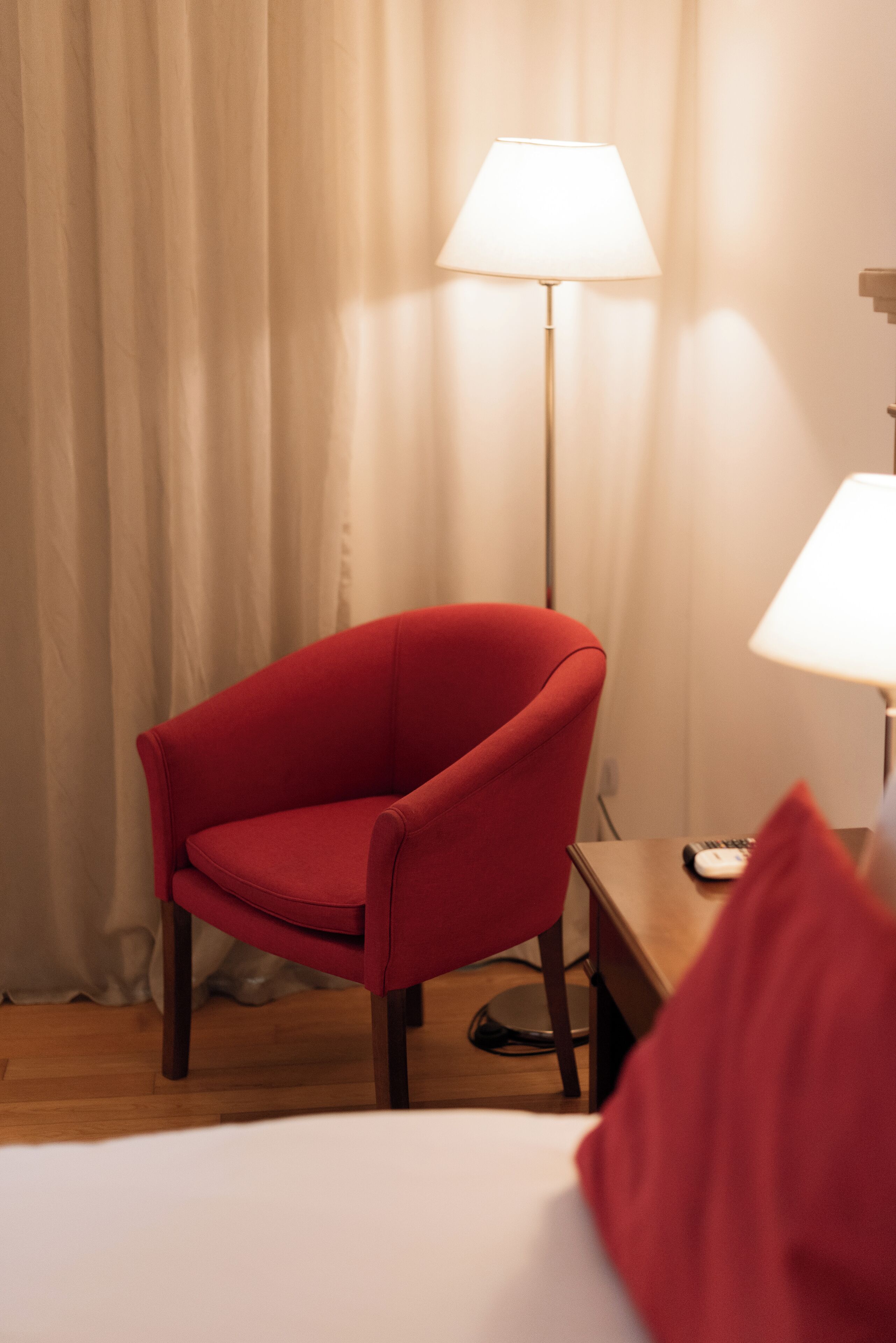 A hotel room with a red armchair, a bedside table with a lamp, and a bed.