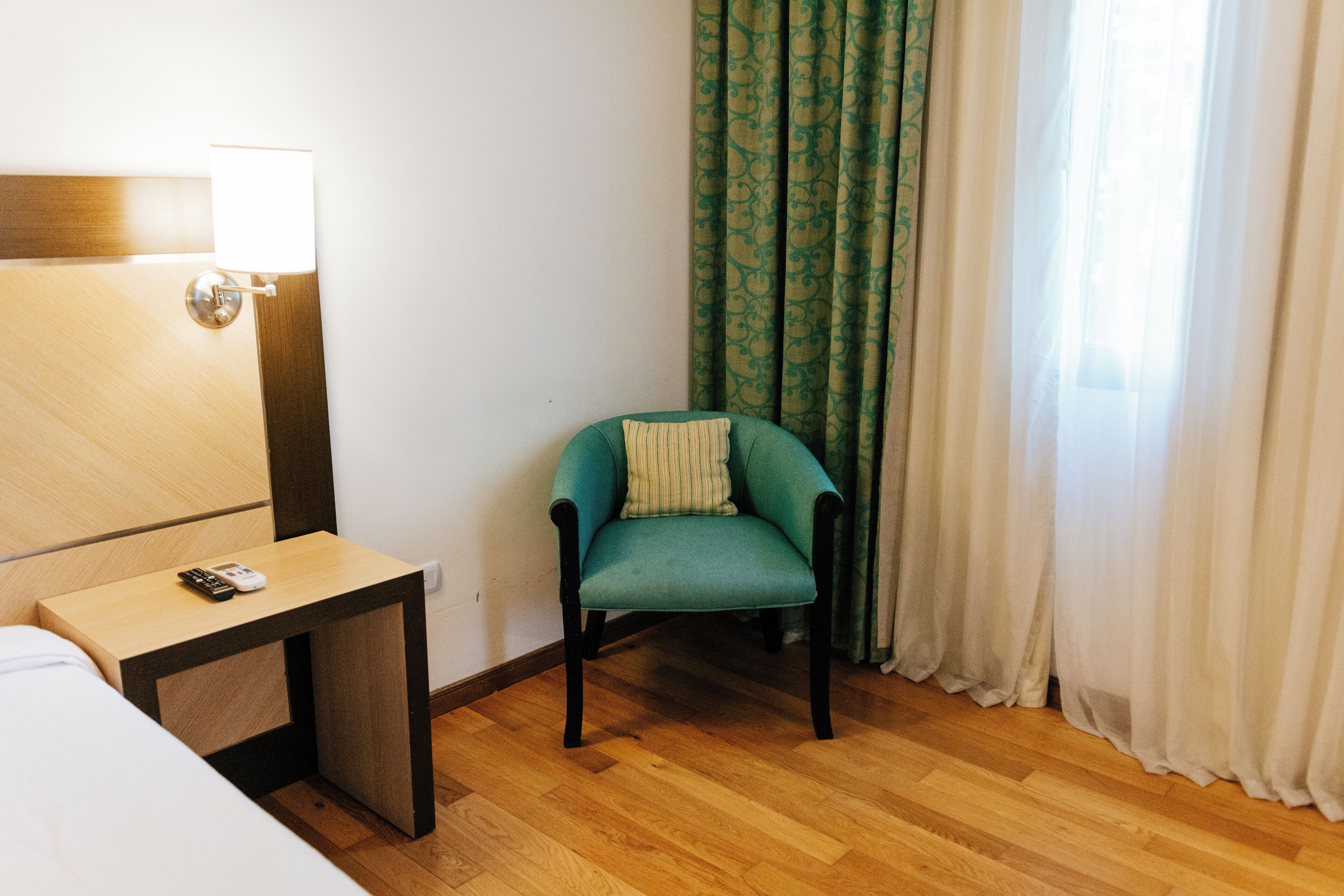 Une chambre d’hôtel avec un plancher en bois, un lit, une petite table avec une télécommande, un fauteuil turquoise agrémenté d’un coussin et une fenêtre avec des rideaux verts.