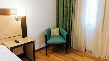 Une chambre d’hôtel avec un plancher en bois, un lit, une petite table avec une télécommande, un fauteuil turquoise agrémenté d’un coussin et une fenêtre avec des rideaux verts.