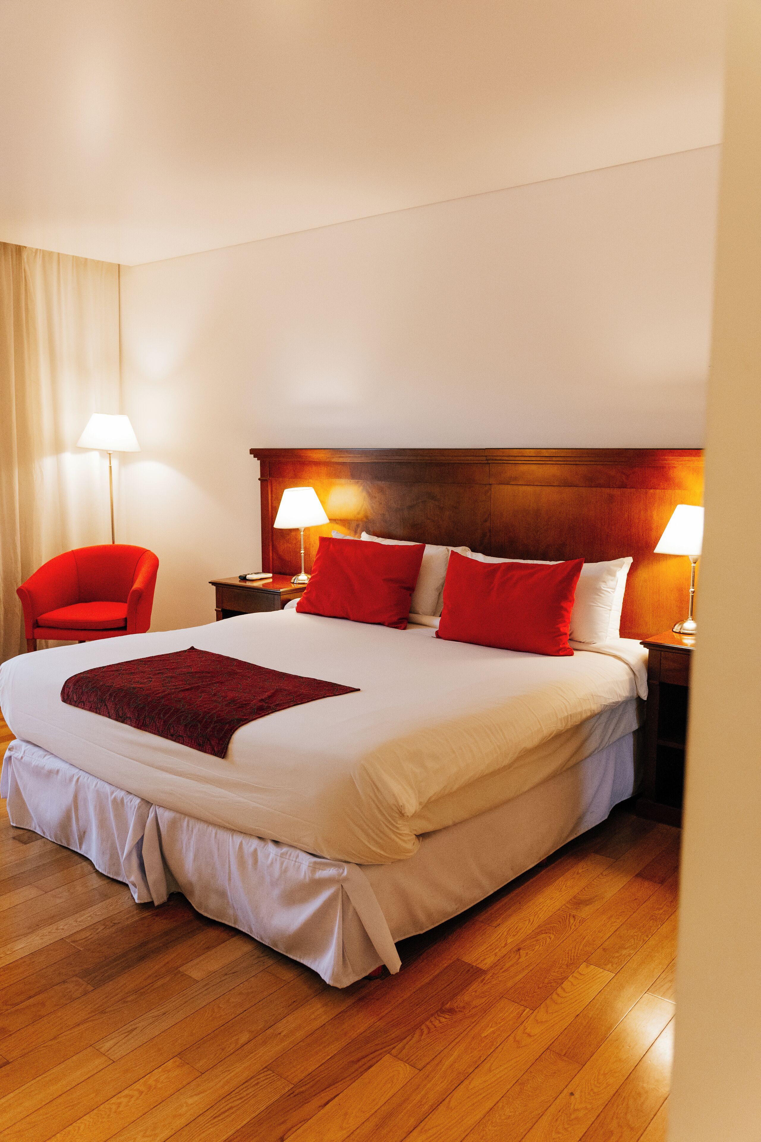 A neatly made bed with red and white pillows, a wooden headboard, and a red chair beside it.