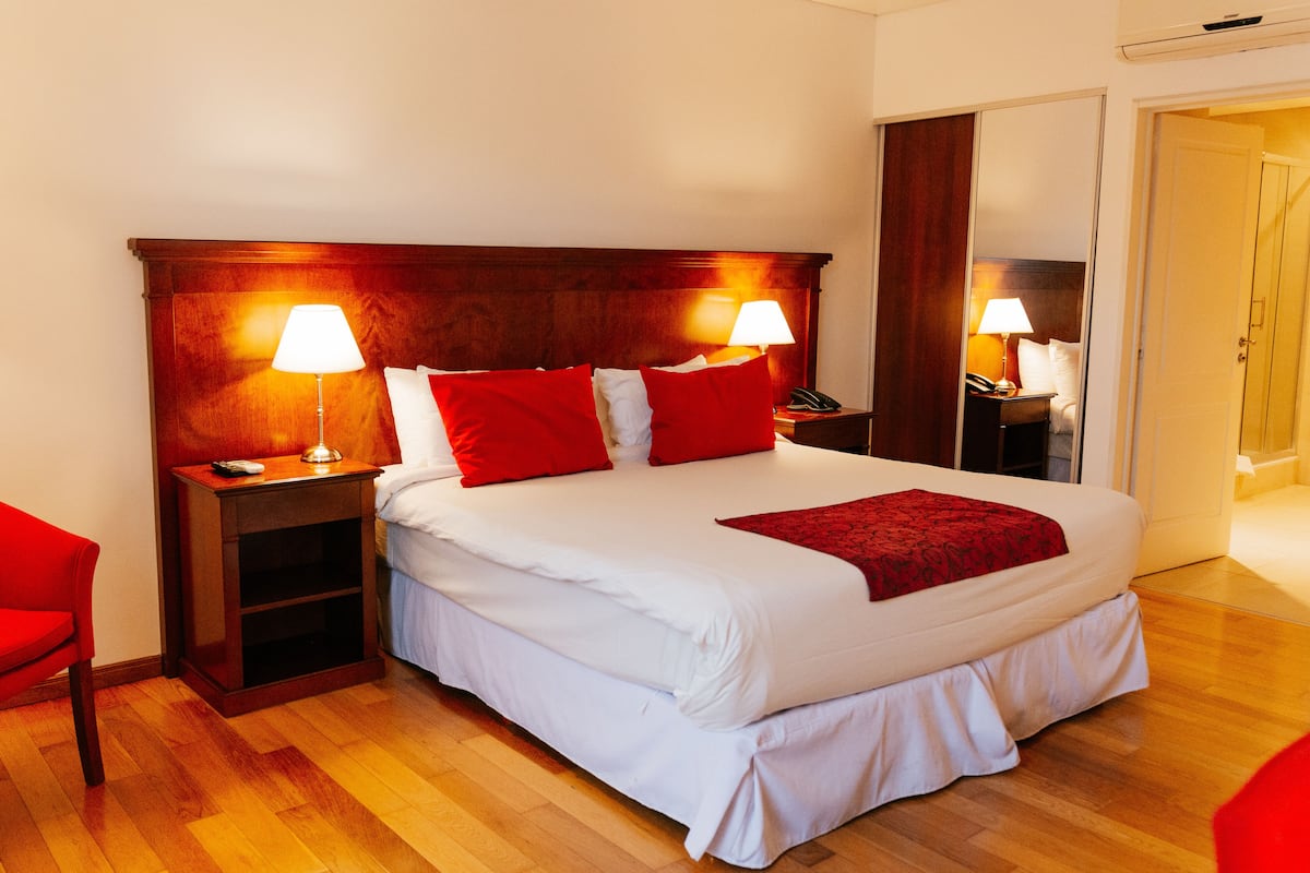 Una cama bien hecha con sábanas blancas y almohadas rojas, dos lámparas de noche, una mesita de madera y una silla roja.