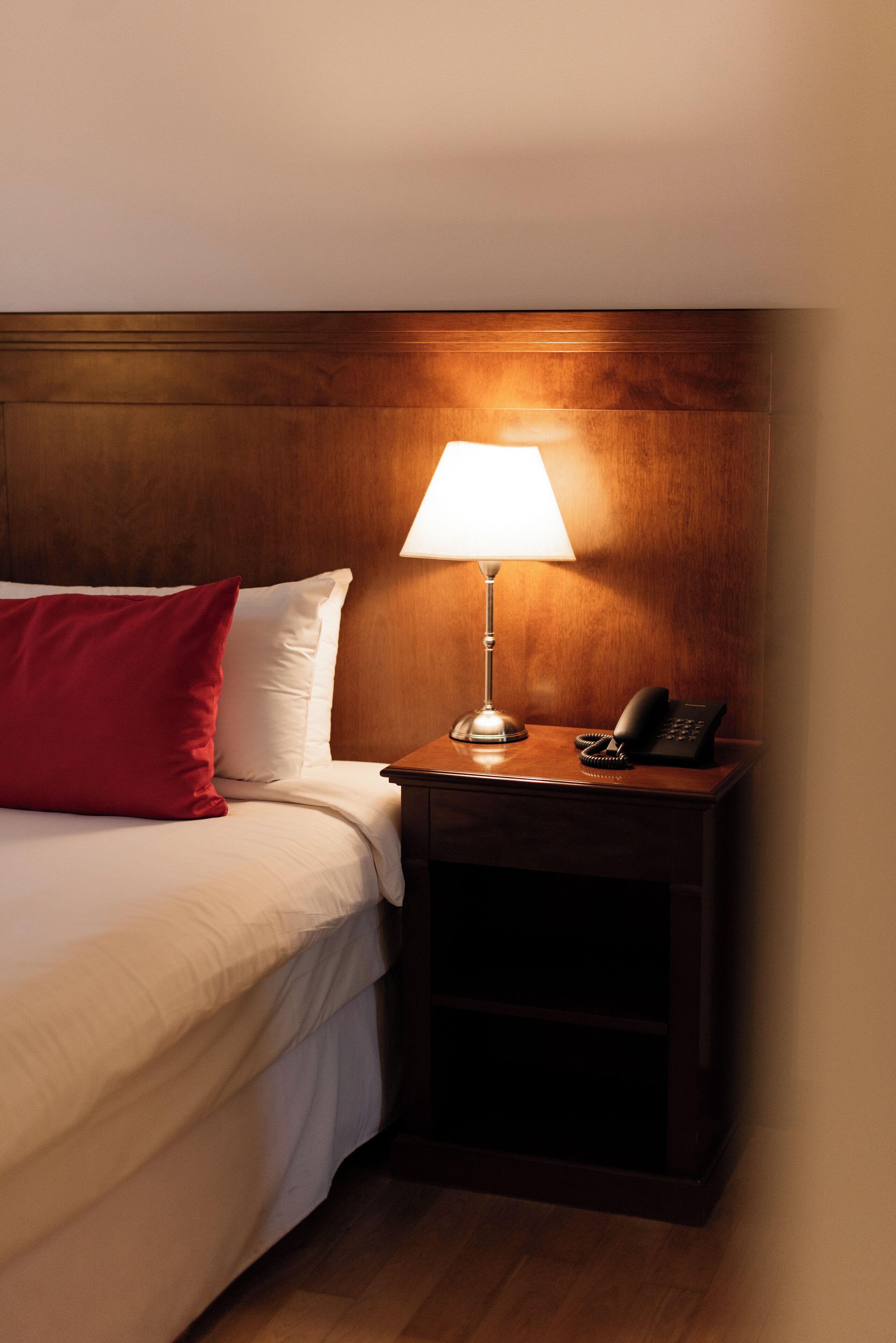 Une chambre d’hôtel avec un lit, une table de chevet avec une lampe, un téléphone et un oreiller rouge.