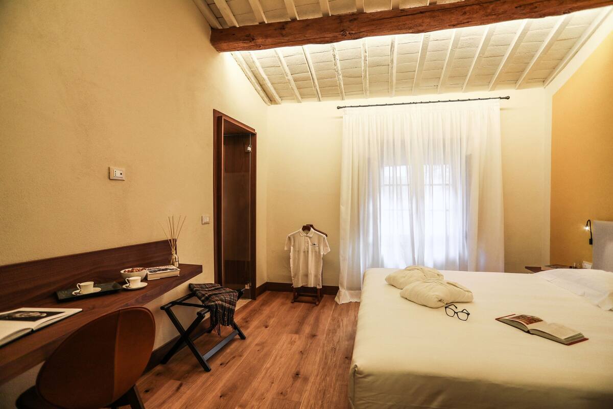 A hotel room with a wooden desk, a bed with pillows and glasses, a hanging robe, and a window with curtains.