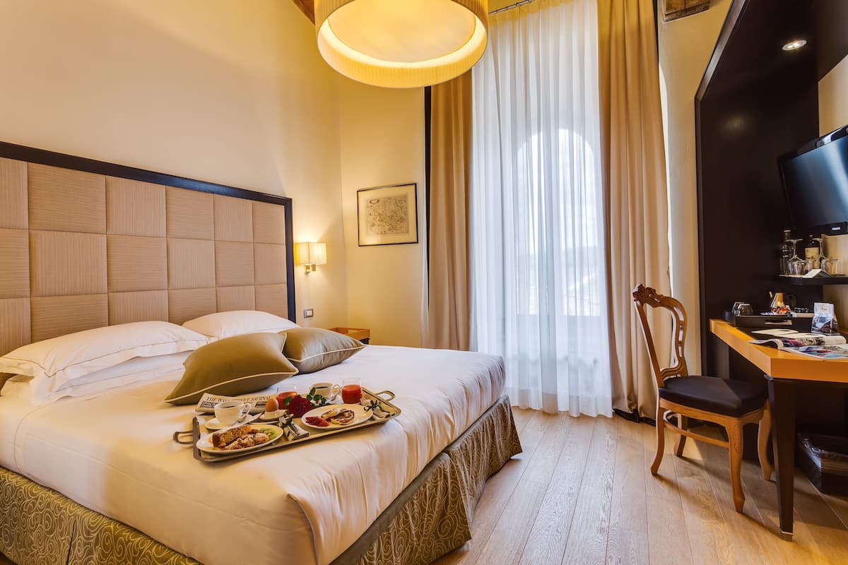 A hotel room with a bed, breakfast tray, a desk with a chair, a television, and a window with curtains.