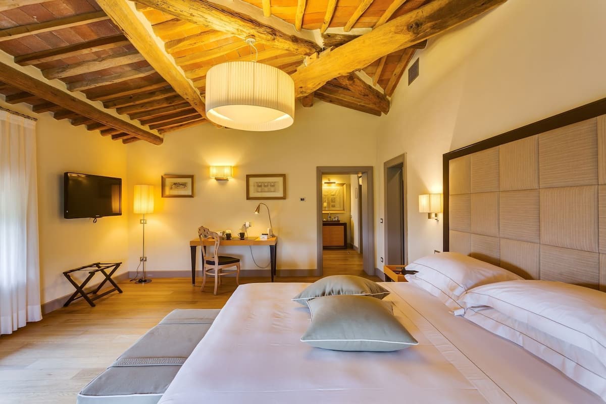 A hotel room with a wooden ceiling, a bed with pillows, a desk with a chair, a TV, and wall-mounted lamps.