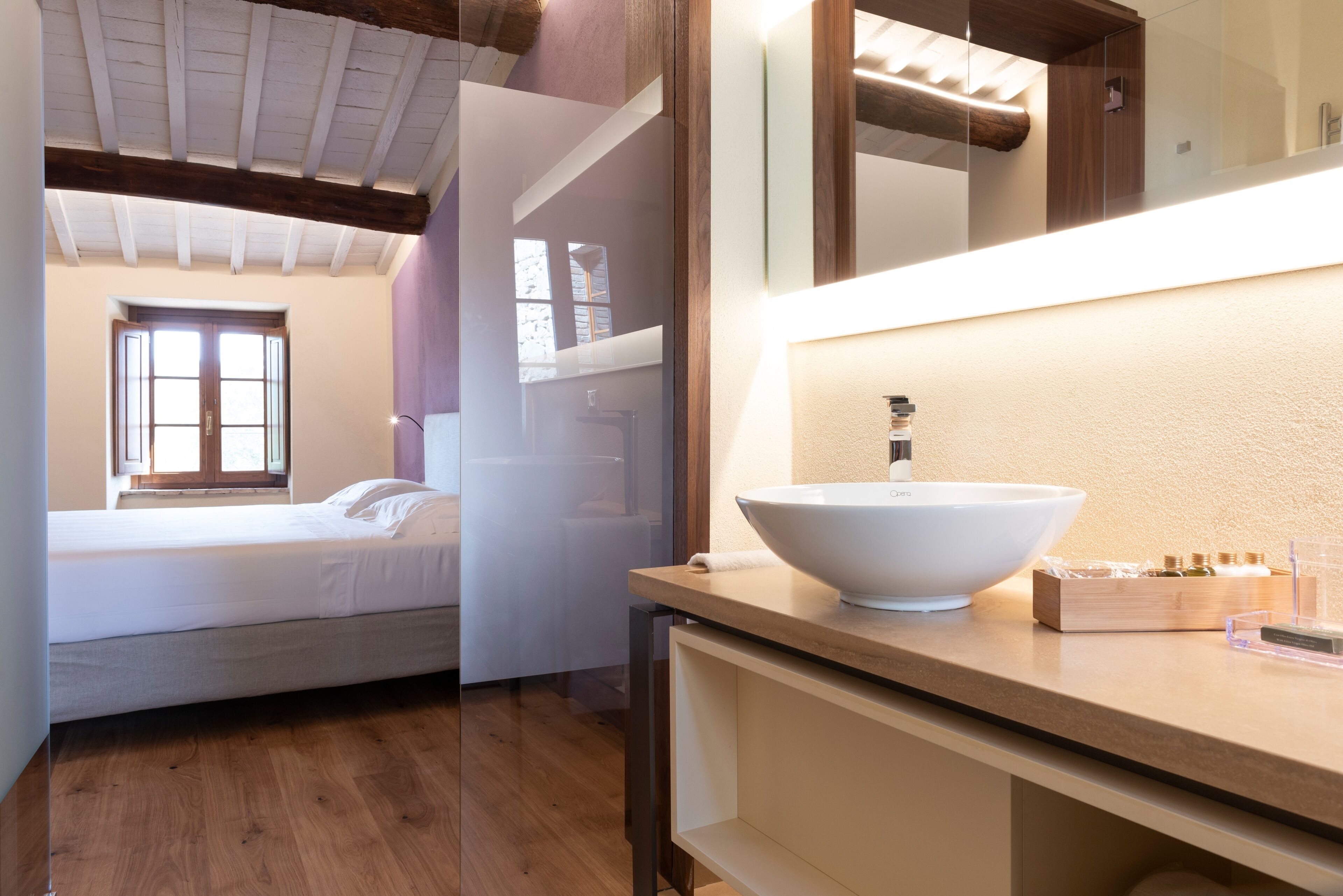 A modern bedroom with a wooden ceiling, a large window, a bed, and a bathroom with a large sink and toiletries.