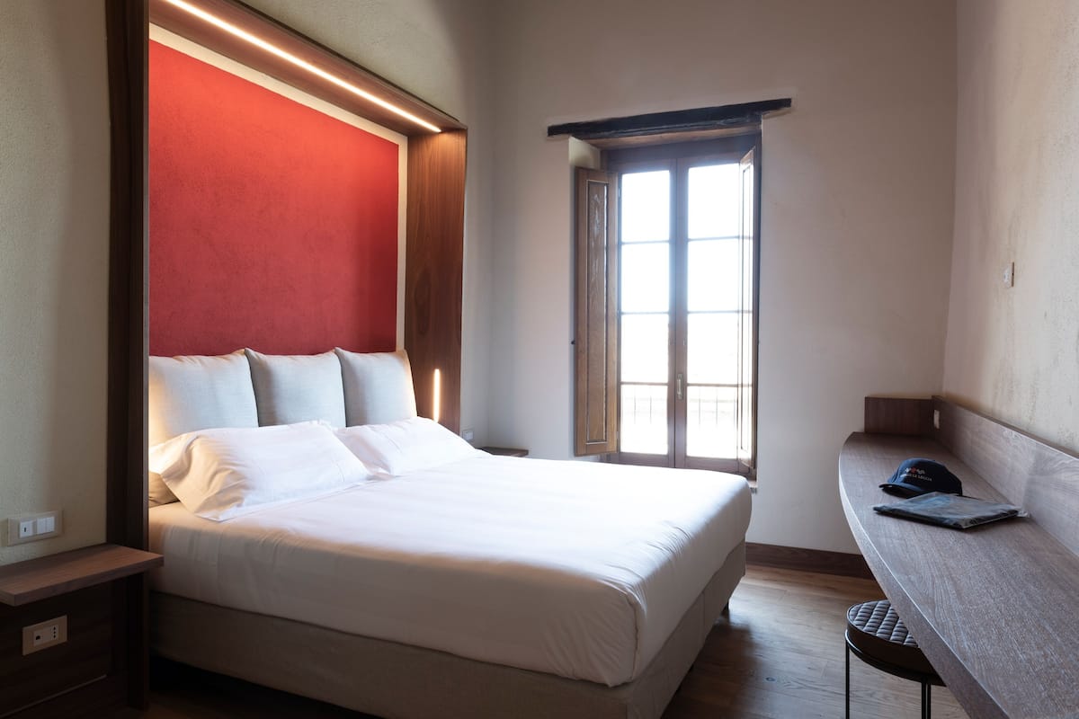 A hotel room with a bed, a desk, a window, and a red accent wall.