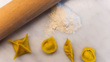Een marmeren oppervlak met een deegroller, bloem en drie gele pasta vormen.